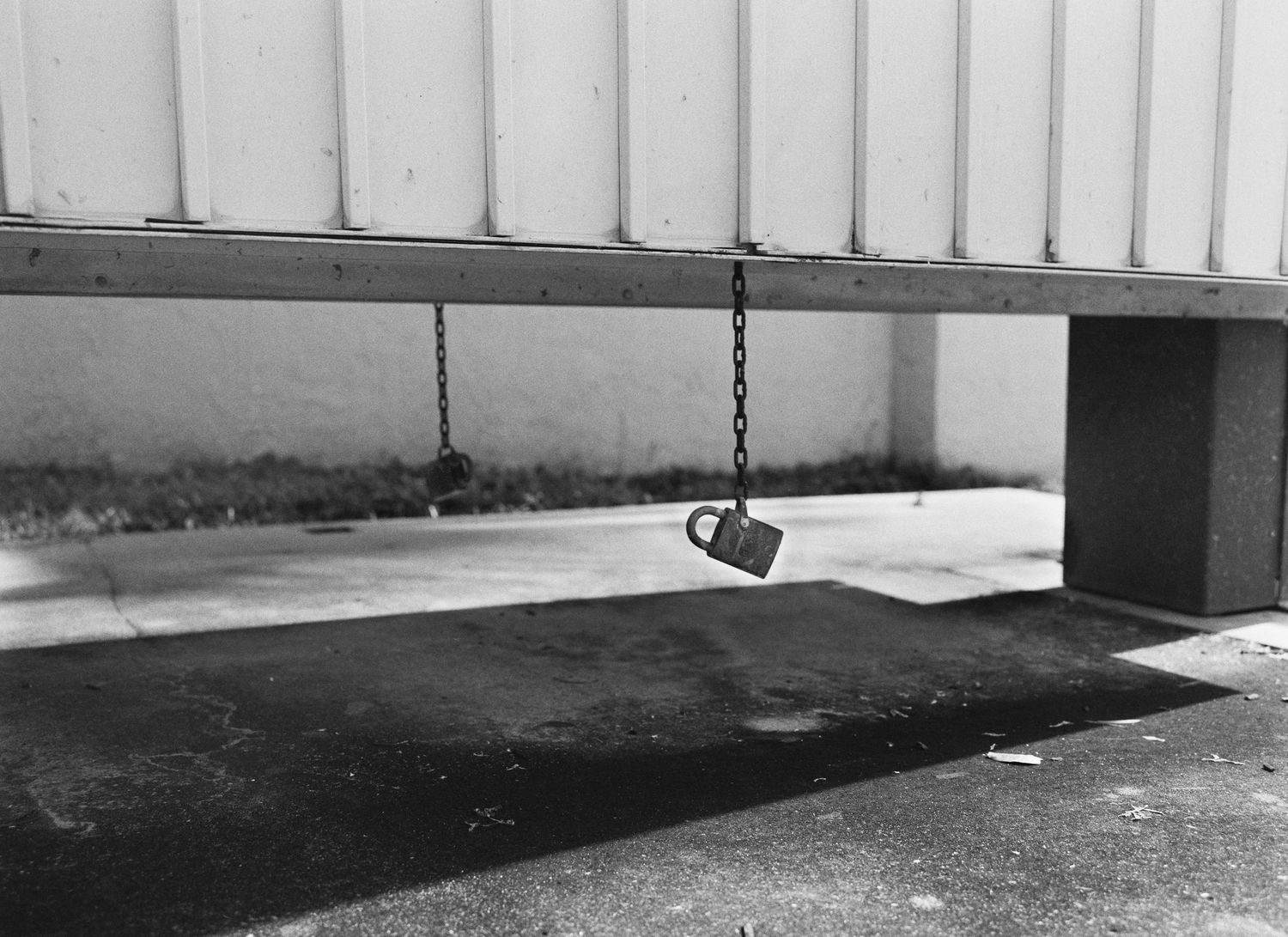 Black and white photo of a padlock hanging on a chain beneath a metal garage door on a concrete surface.
