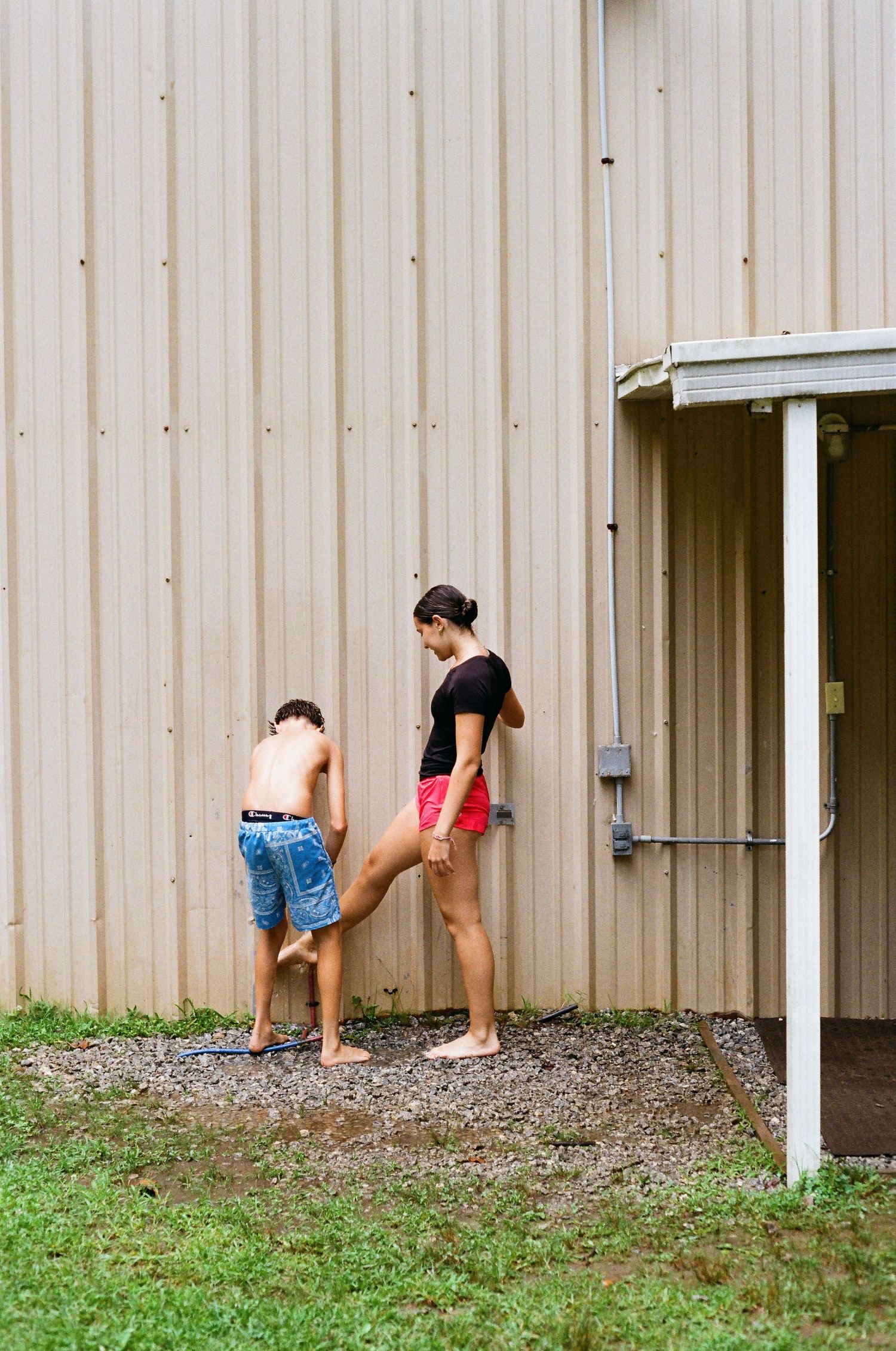 A woman in red shorts and a child in blue jeans stand barefoot outside a metal building on gravel.