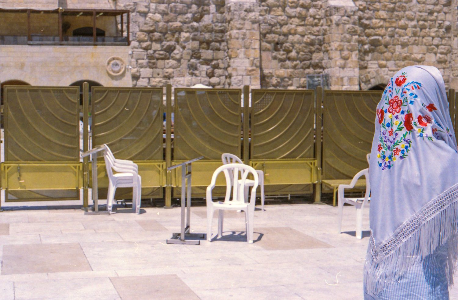 Woman in embroidered shawl praying at the Western Wall plaza with white chairs and green dividers.