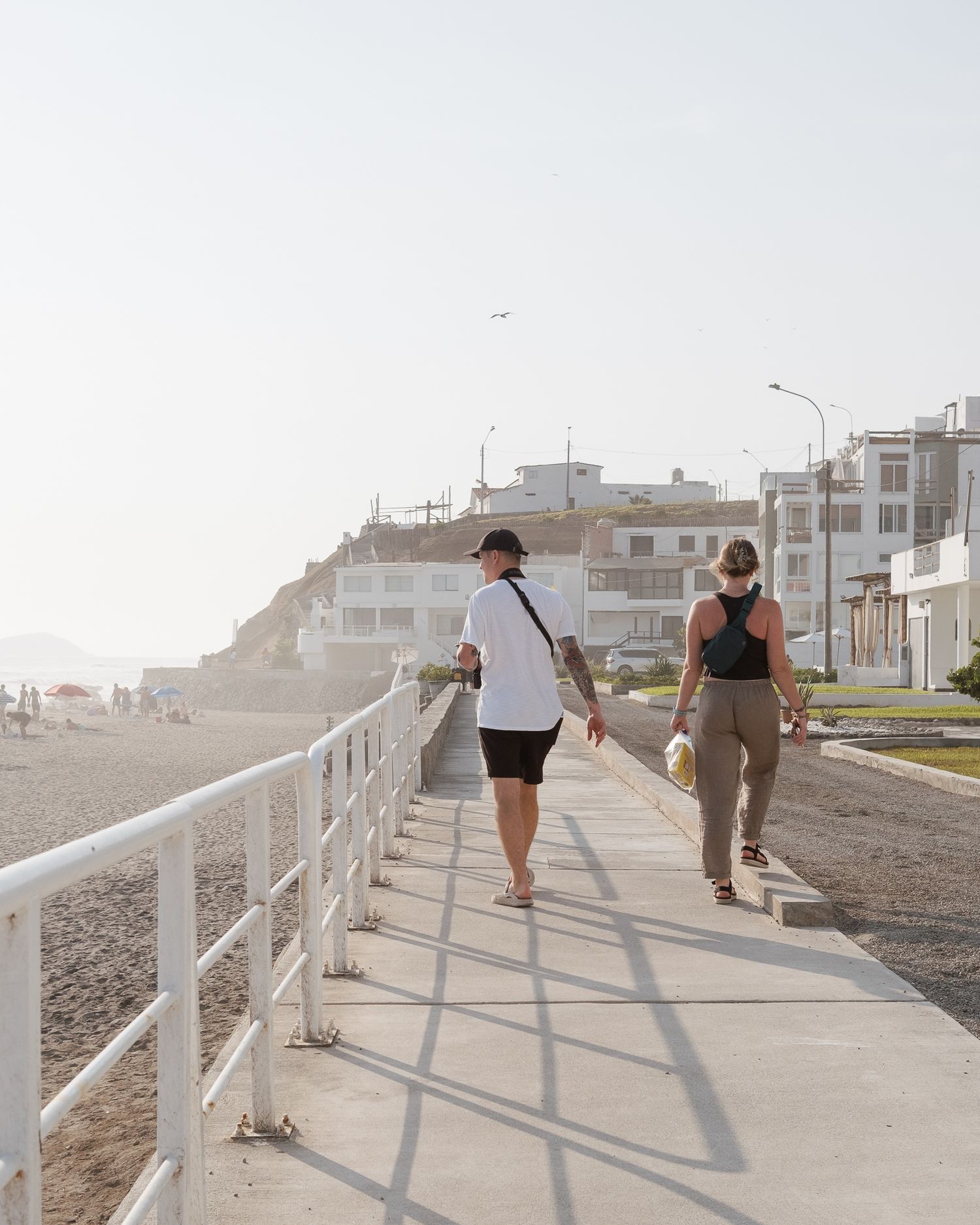 Two people walking along a coastal promenade with white railings and buildings in the background on a sunny day.