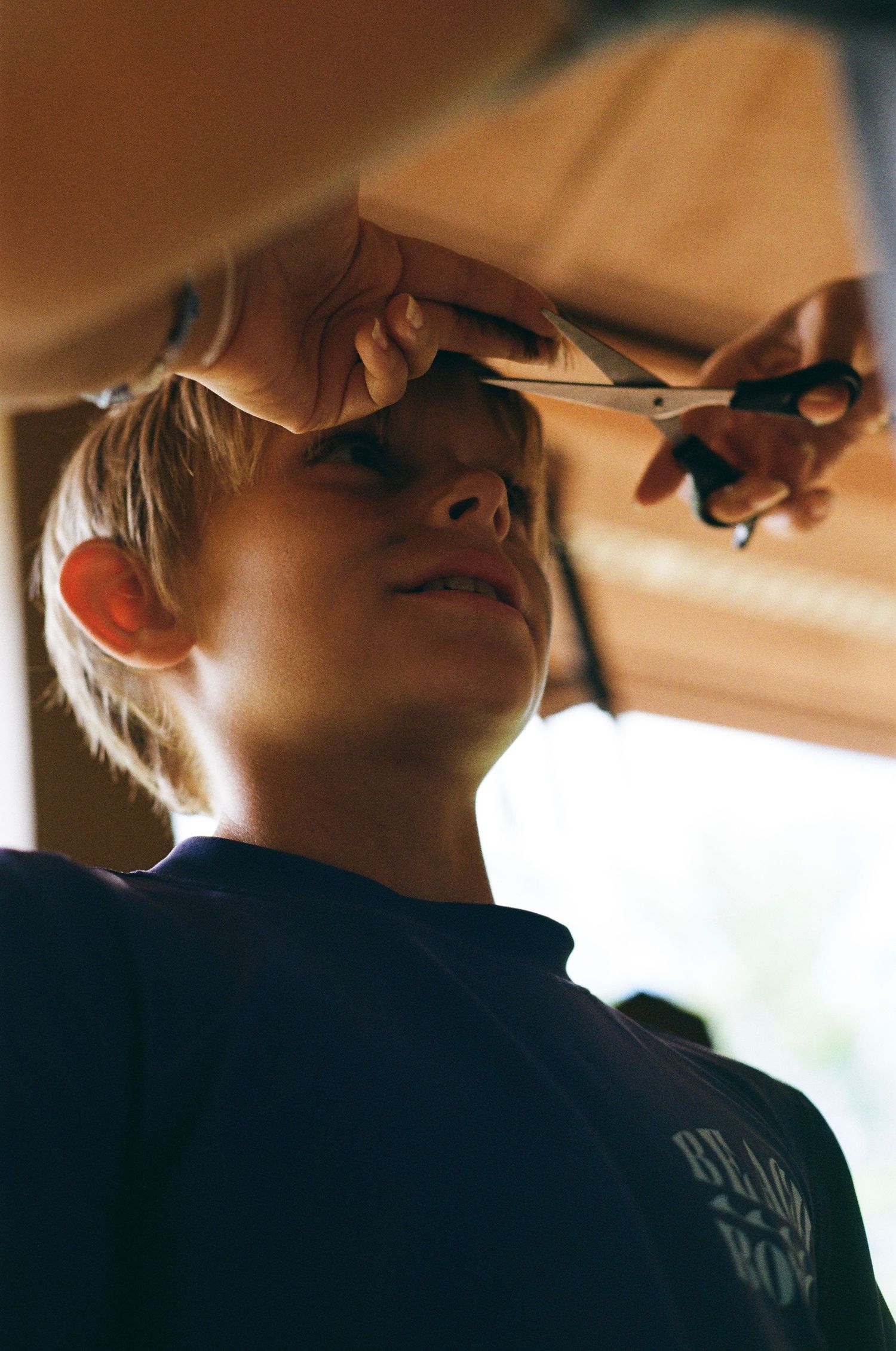 A young blonde boy getting a haircut with scissors held above his head, wearing a dark blue shirt.