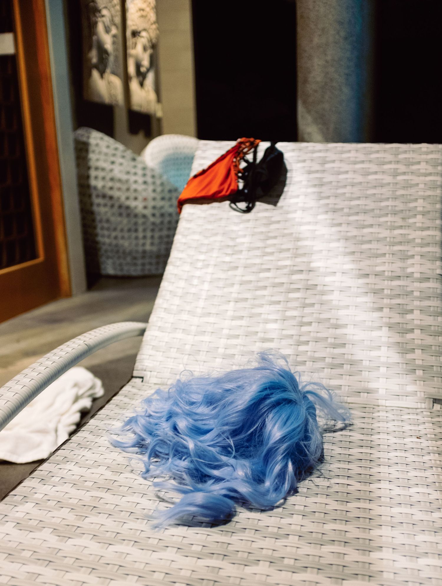 Blue curly wig and red bikini top resting on a white woven lounge chair indoors.