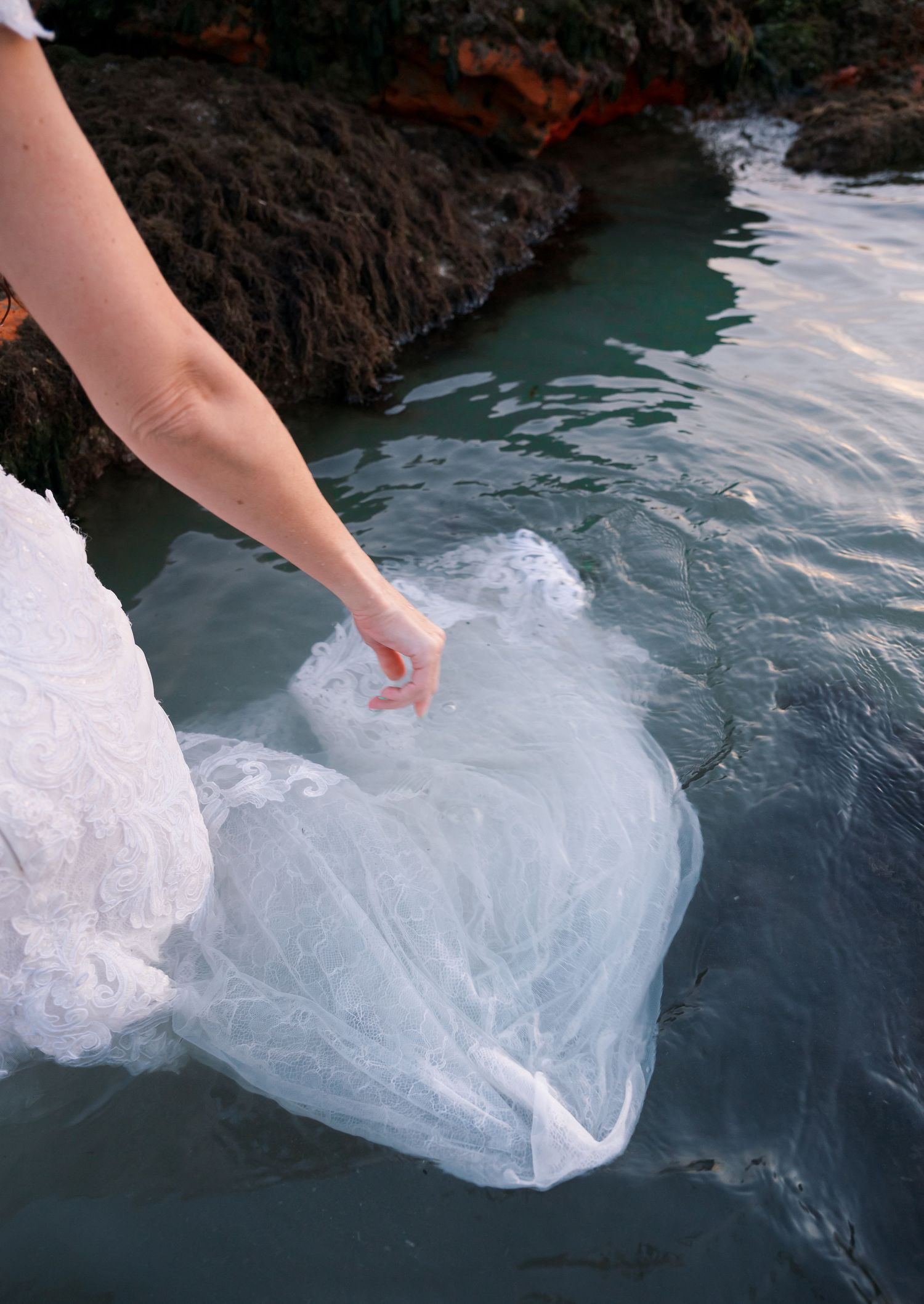 Hand releases plastic bag containing water into turquoise ocean water near rocky coastline.