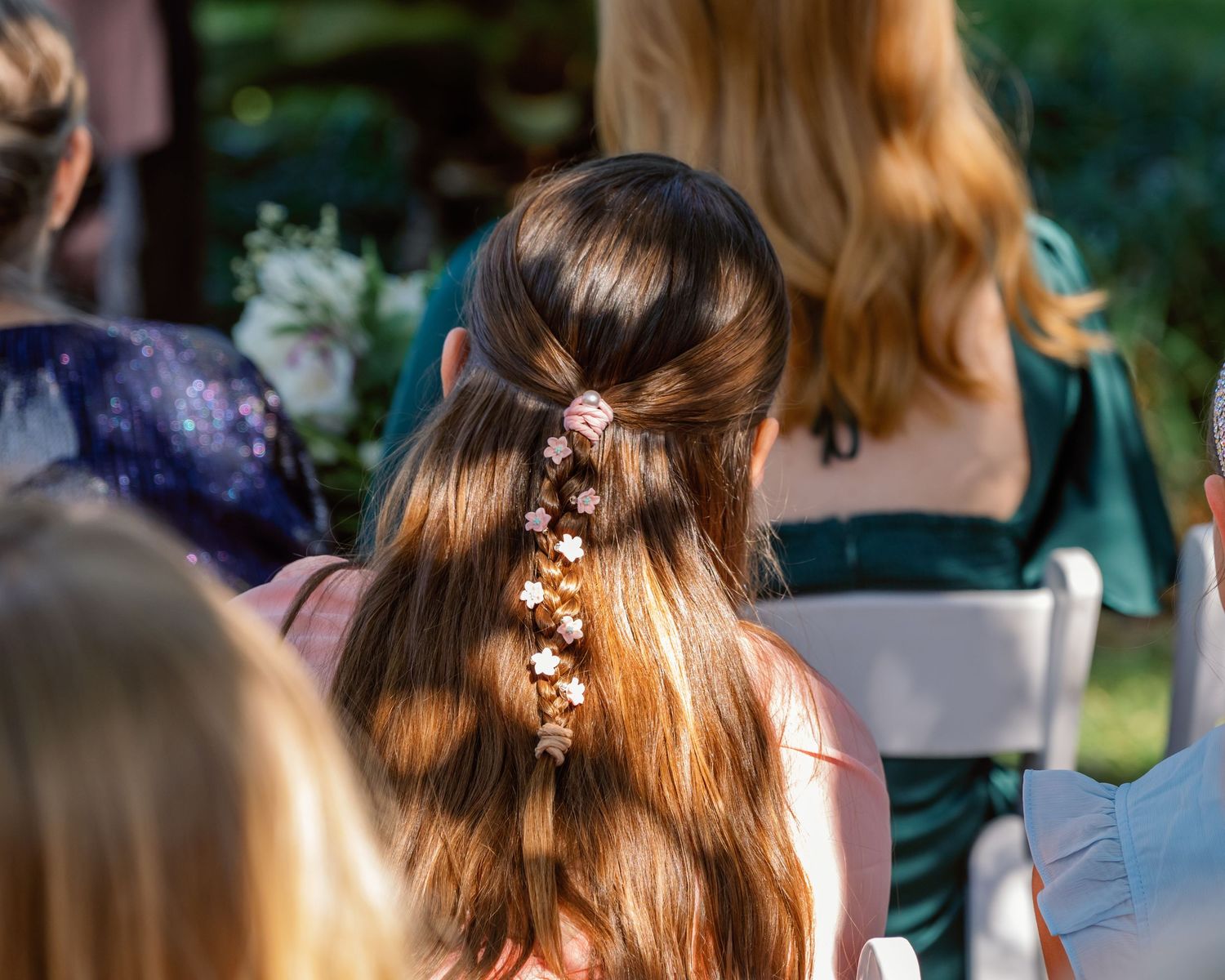 Delicate floral hair accessories adorn a hairstyle with small white flowers and greenery against a blurred outdoor background.