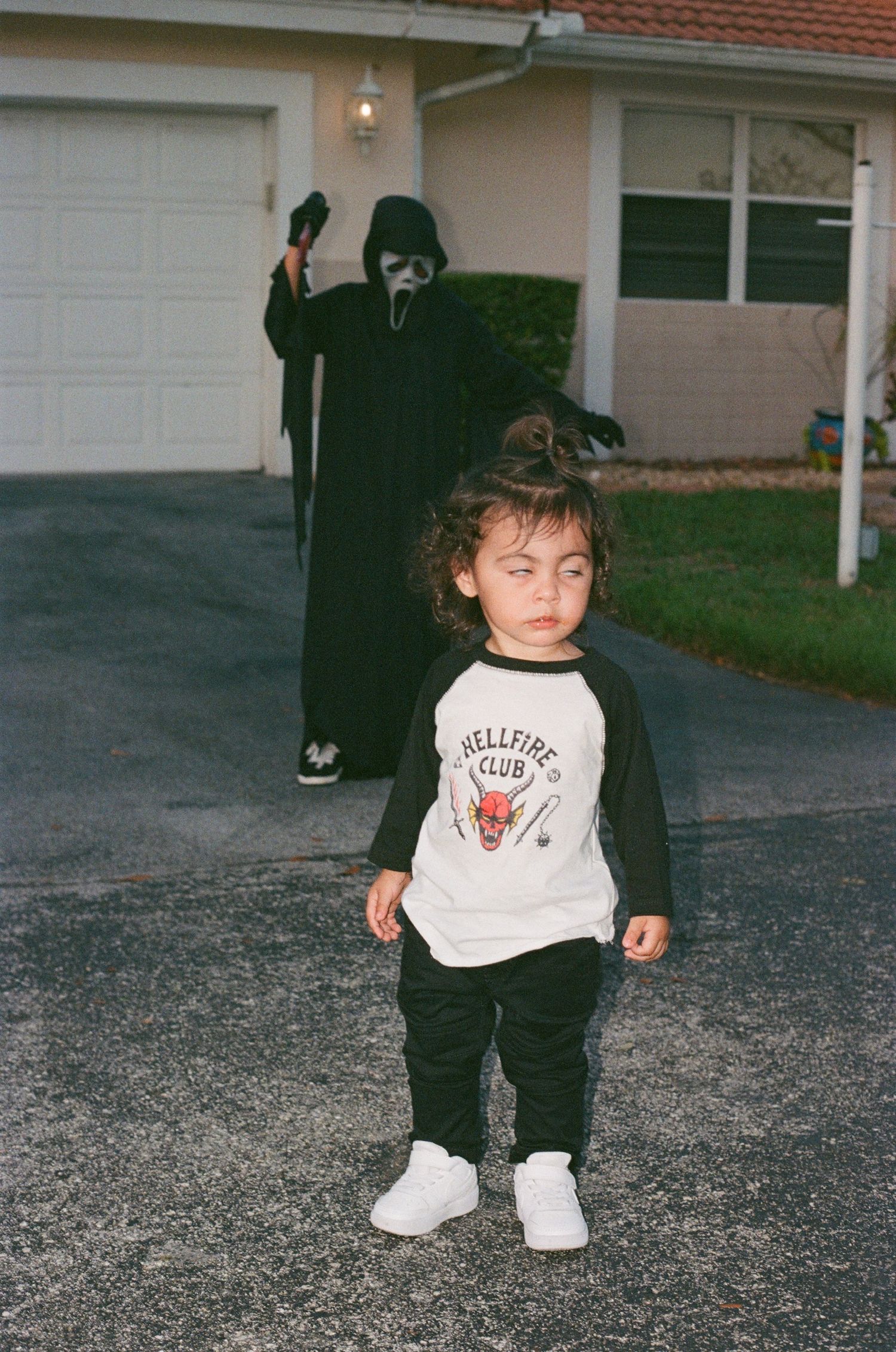 A toddler wearing a white graphic t-shirt and dark pants stands on a gravel driveway near a house.