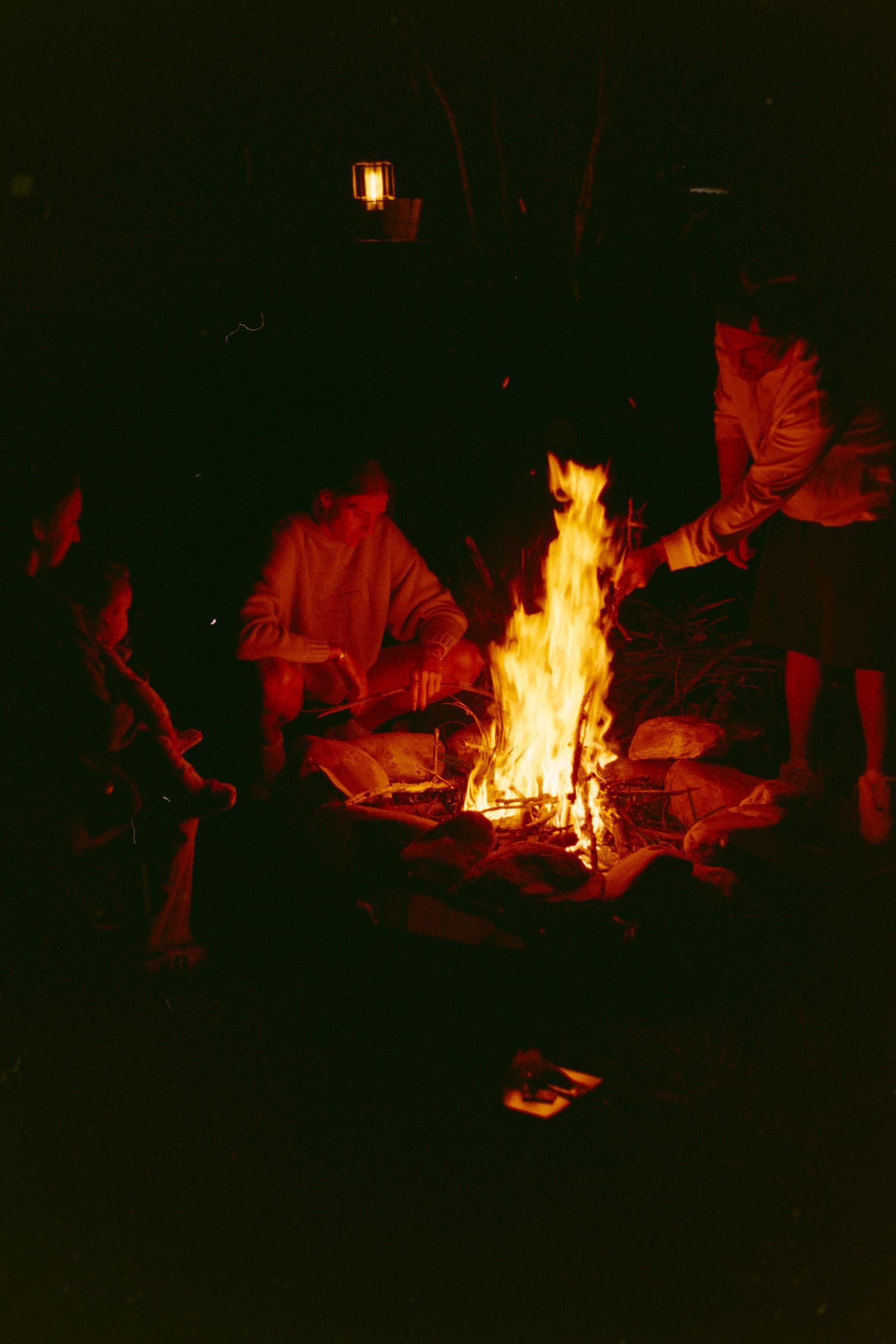 Group of people gathered around a glowing campfire at night, illuminated by warm orange flames in a dark outdoor setting.