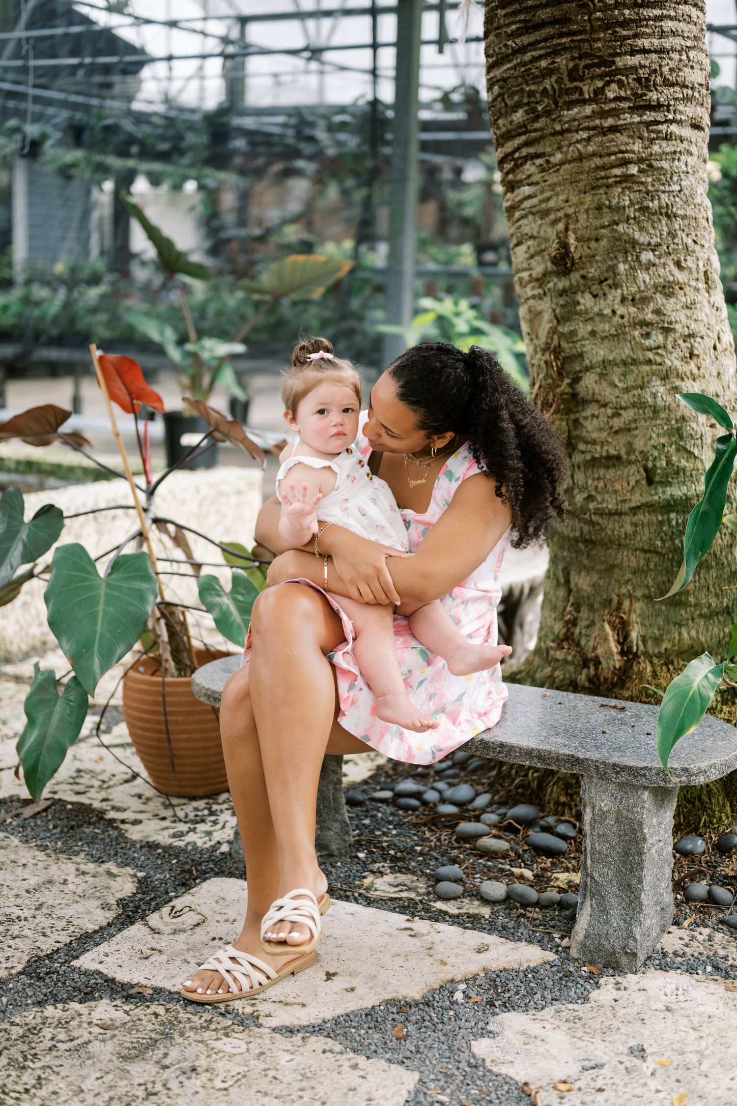 Two people in matching pink dresses share a tender moment while sitting on a stone bench under a palm tree.