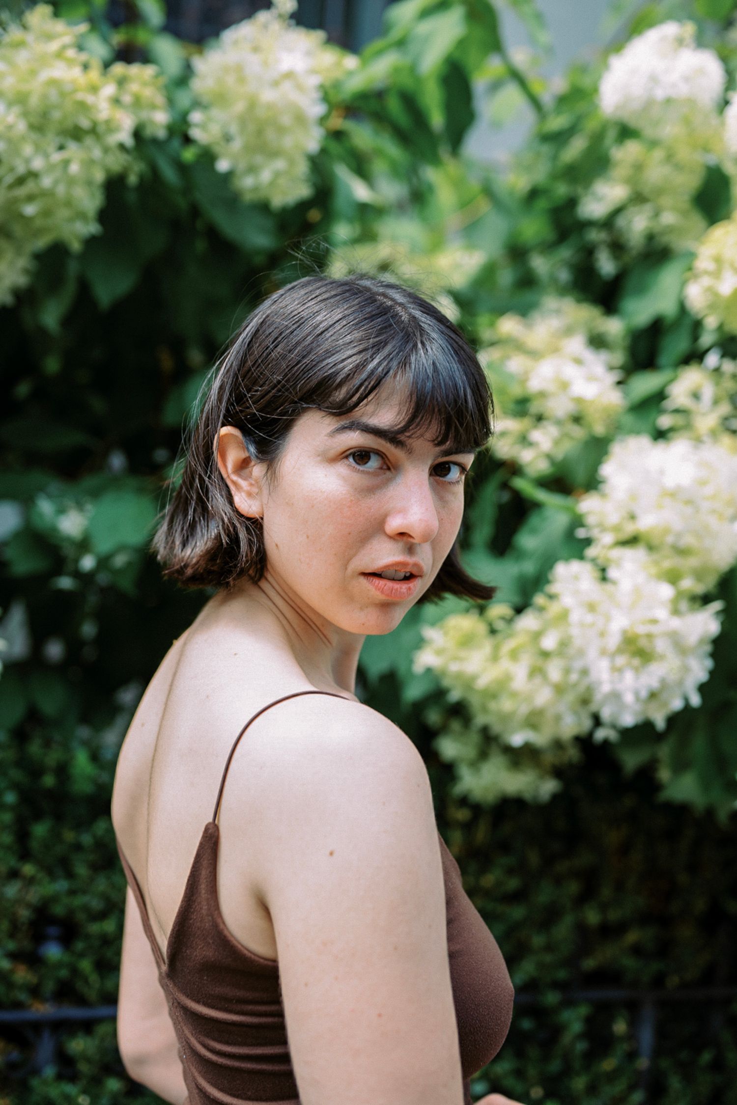 A figure poses against a backdrop of white hydrangea blooms and green foliage in a garden setting.