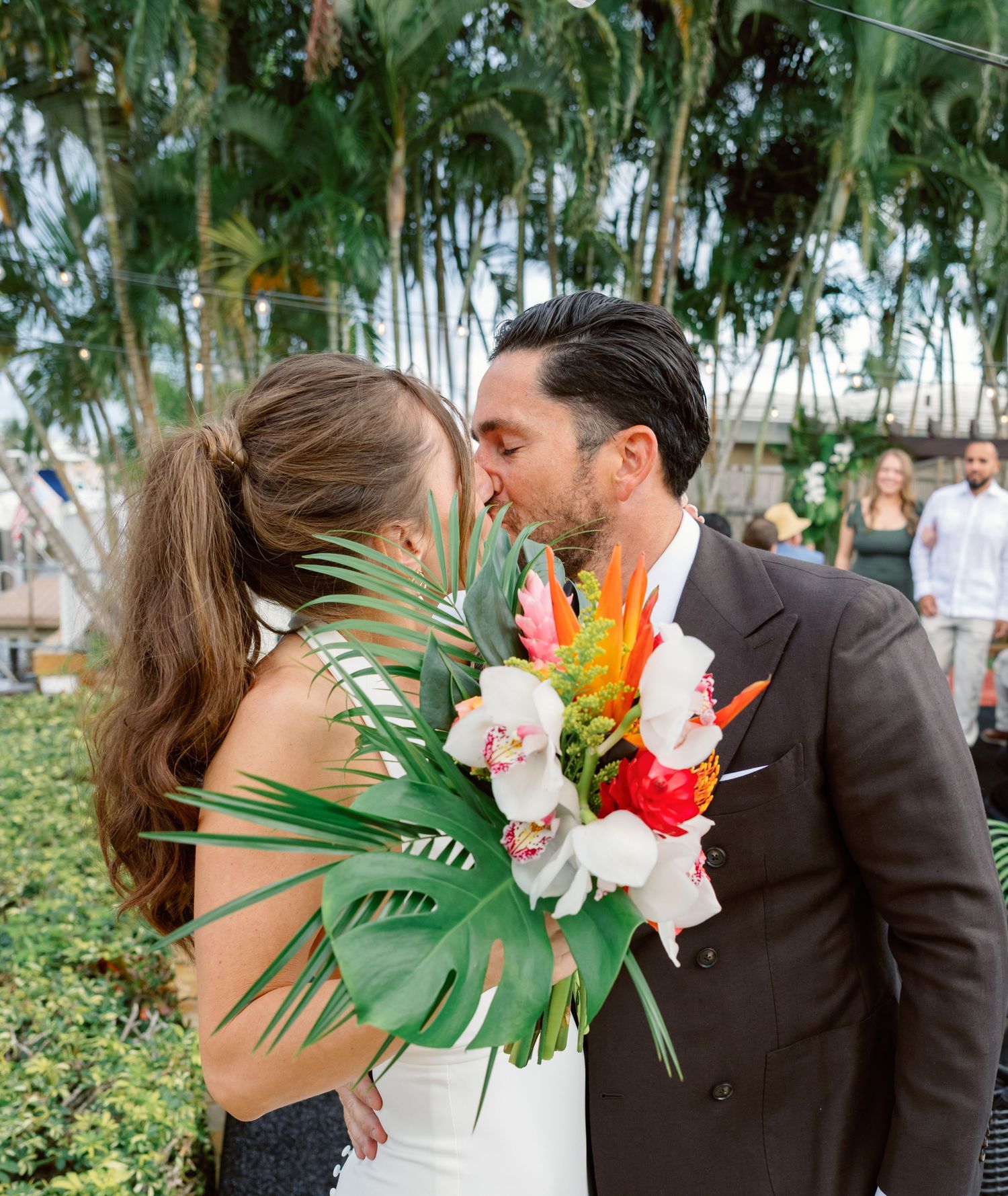 Romantic wedding couple embracing outdoors surrounded by lush tropical greenery and palm trees.