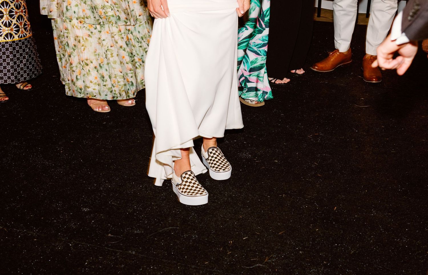 A person wearing a floral dress and white shoes walks across a dark floor in a dimly lit setting.