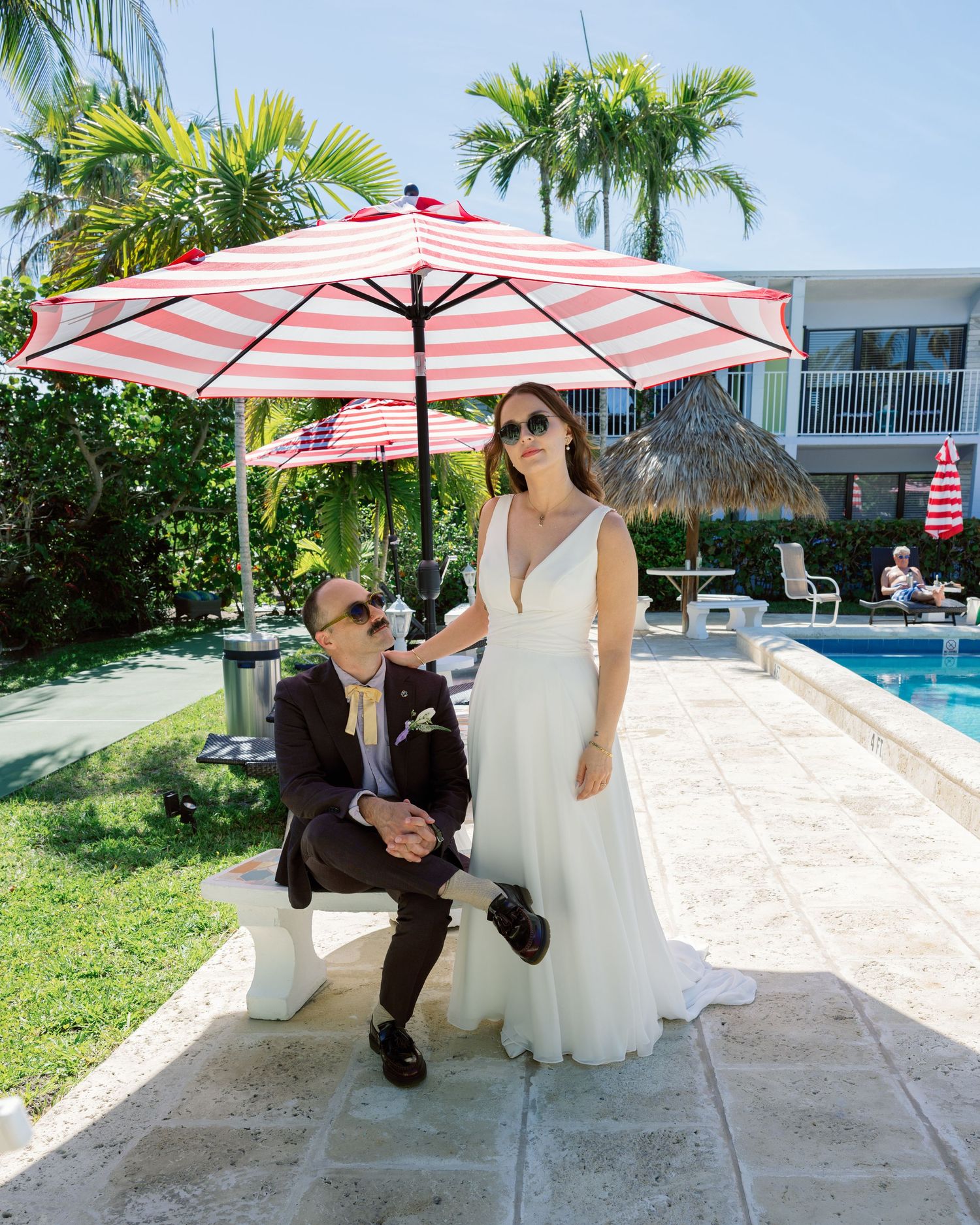 Wedding couple sits under coral striped umbrellas by a poolside on a sunny tropical day.
