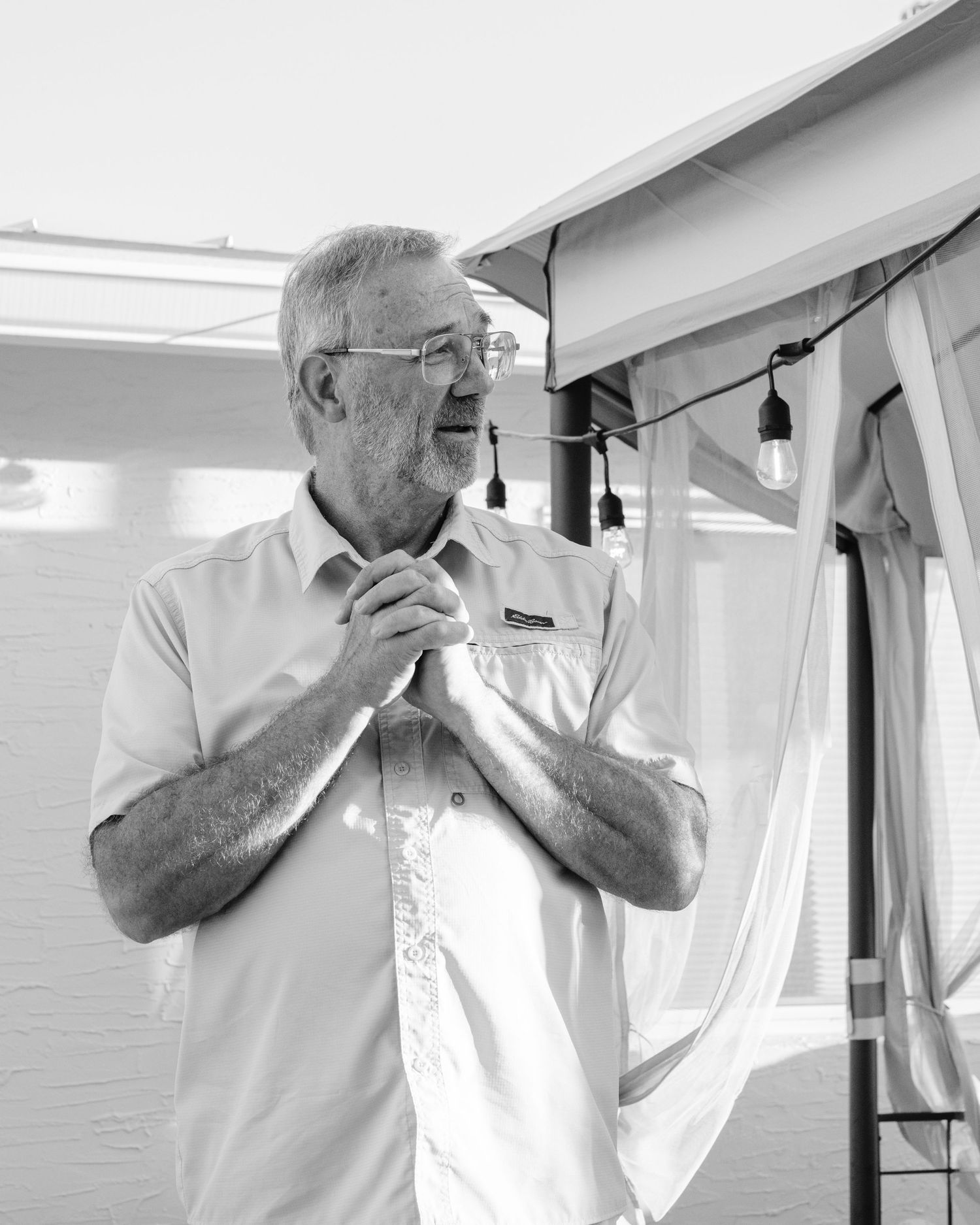 A person in a white shirt stands in an interior space with curtains and overhead lighting, gesturing thoughtfully.