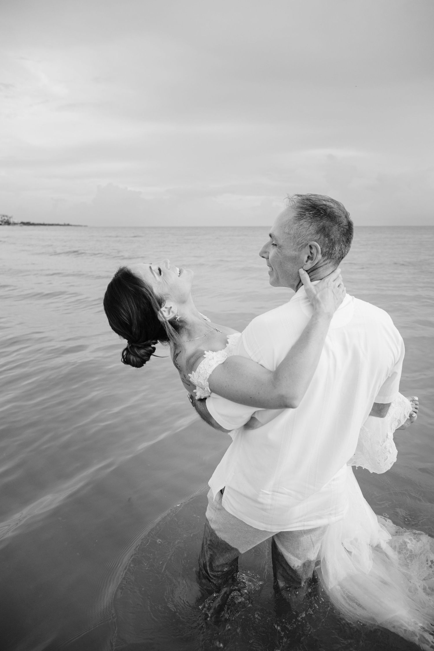 A couple playfully spins in the ocean waves wearing white wedding attire.