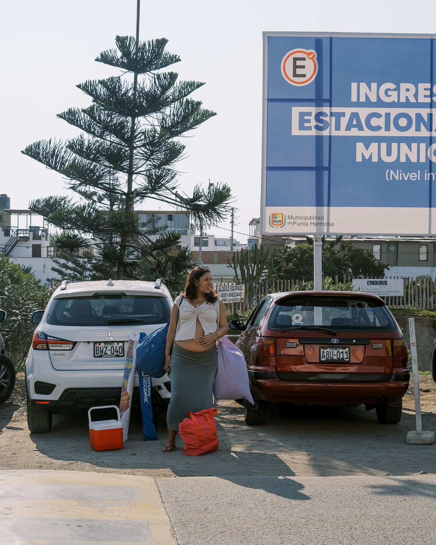 Woman standing between parked cars near a municipal parking entrance sign, holding bags with luggage on the ground.