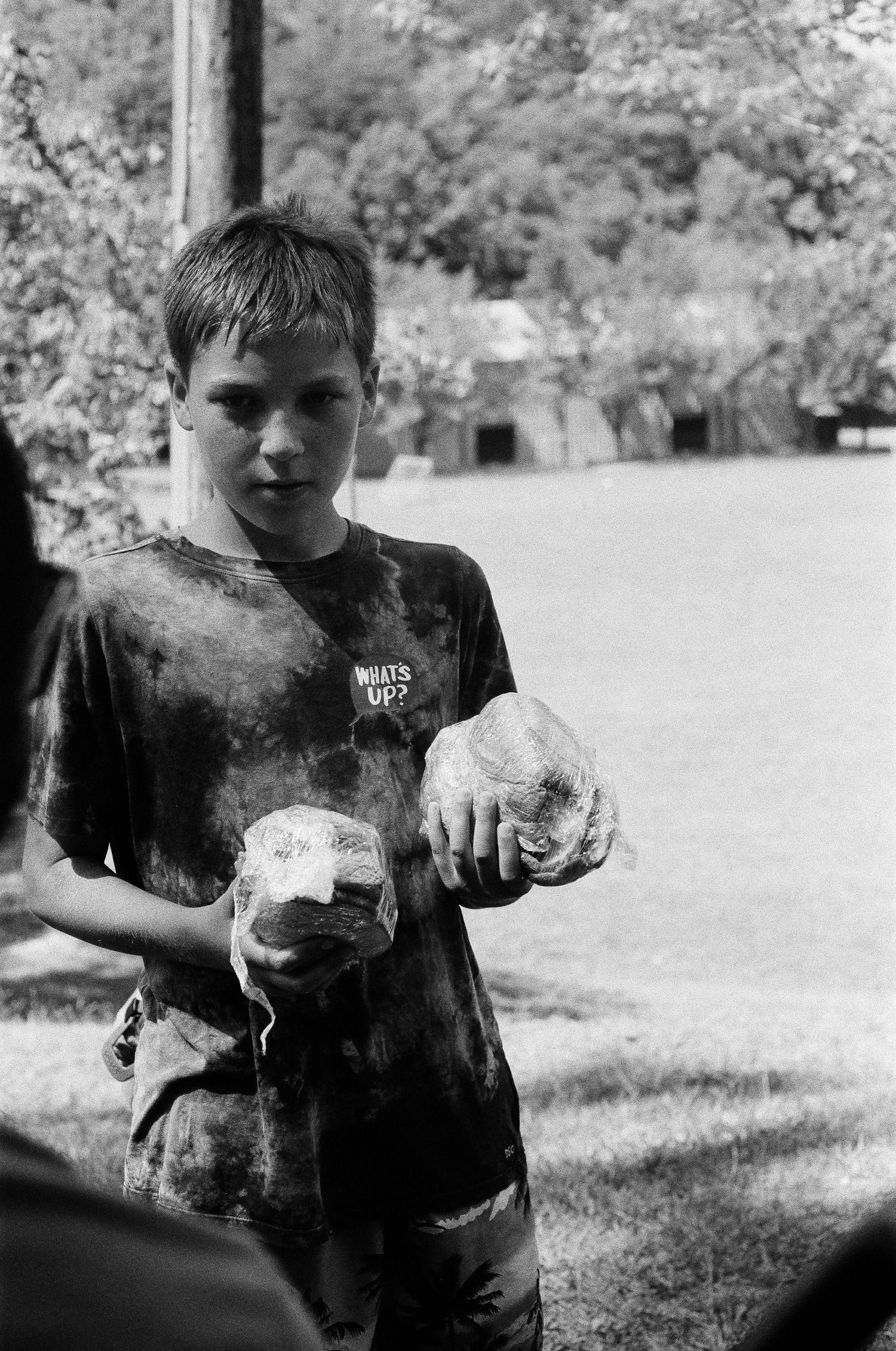 Black and white photo of a young boy in a tie-dye shirt holding two rocks outdoors.