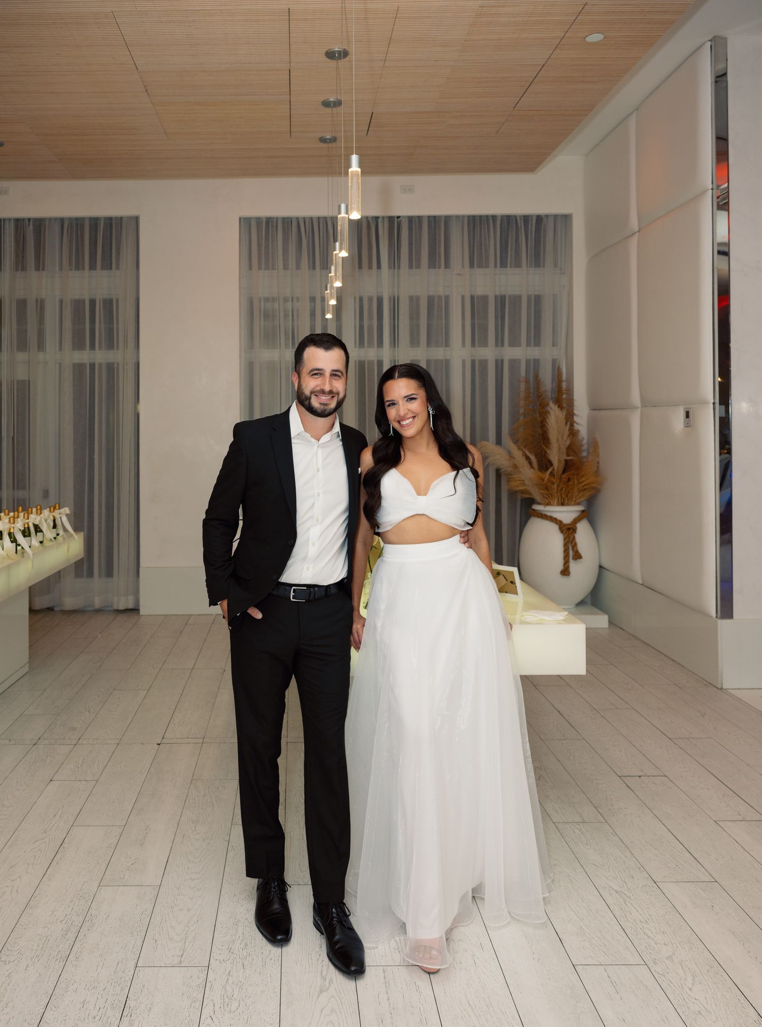 A couple in wedding attire walks hand in hand across a modern white interior space with wood-paneled ceiling.