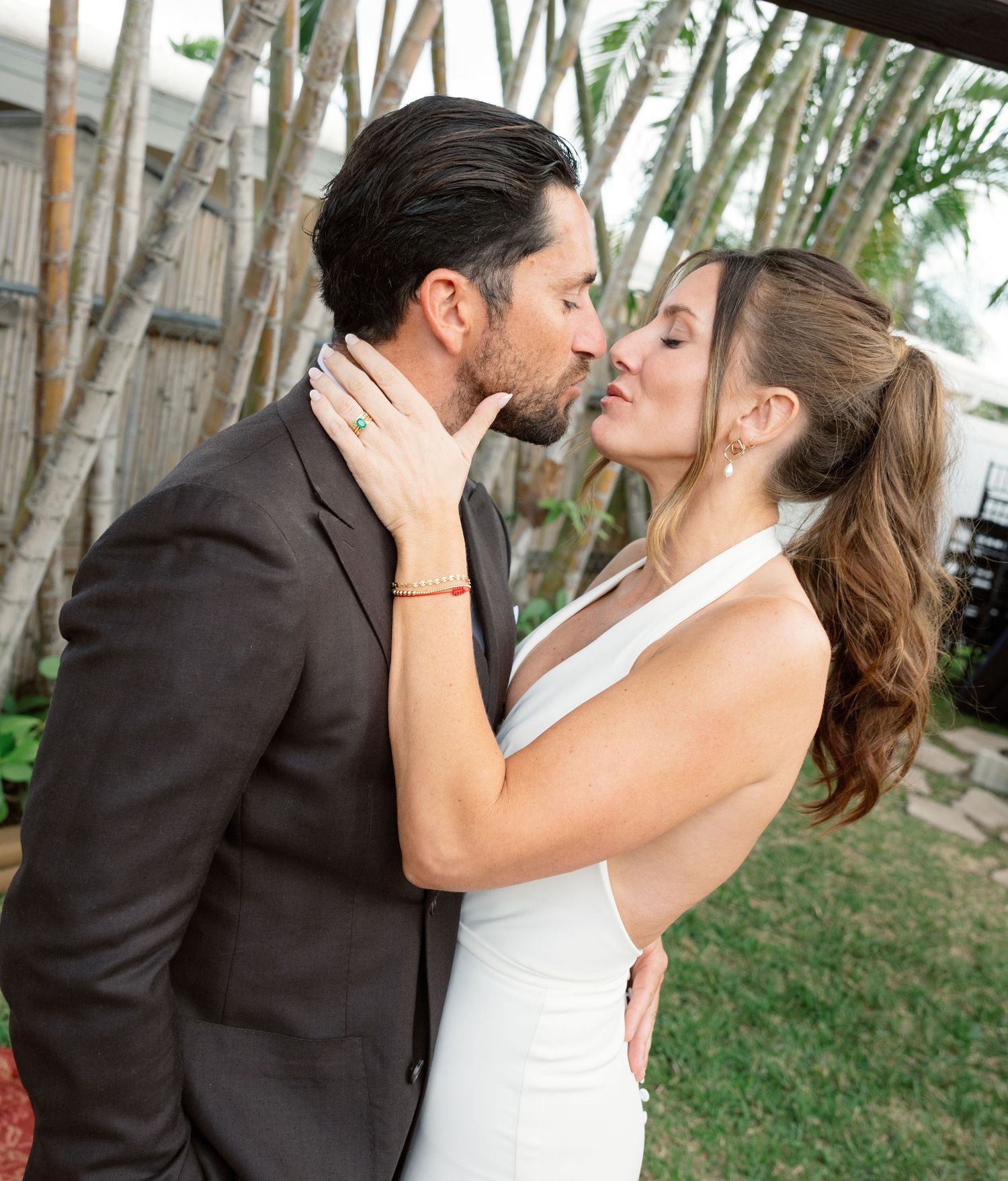 A couple in wedding attire shares a romantic moment outdoors among palm trees and greenery.