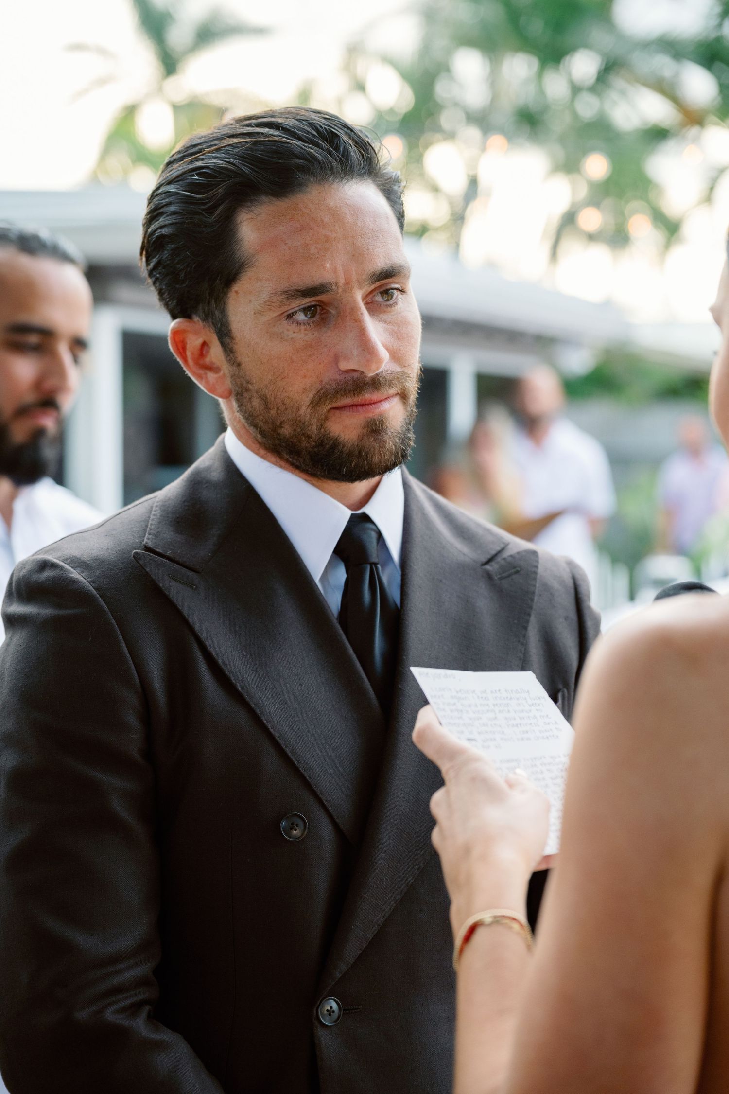 Groom in black suit reading emotional wedding vows during outdoor ceremony.