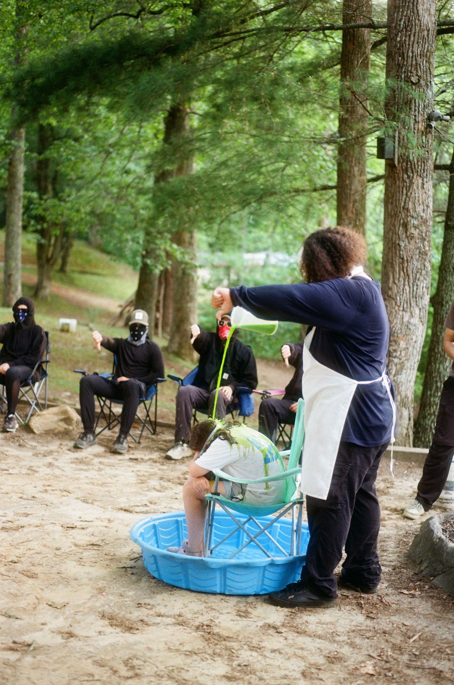 Person in apron holds a fish over a blue kiddie pool outdoors, while masked audience watches in a forest setting.