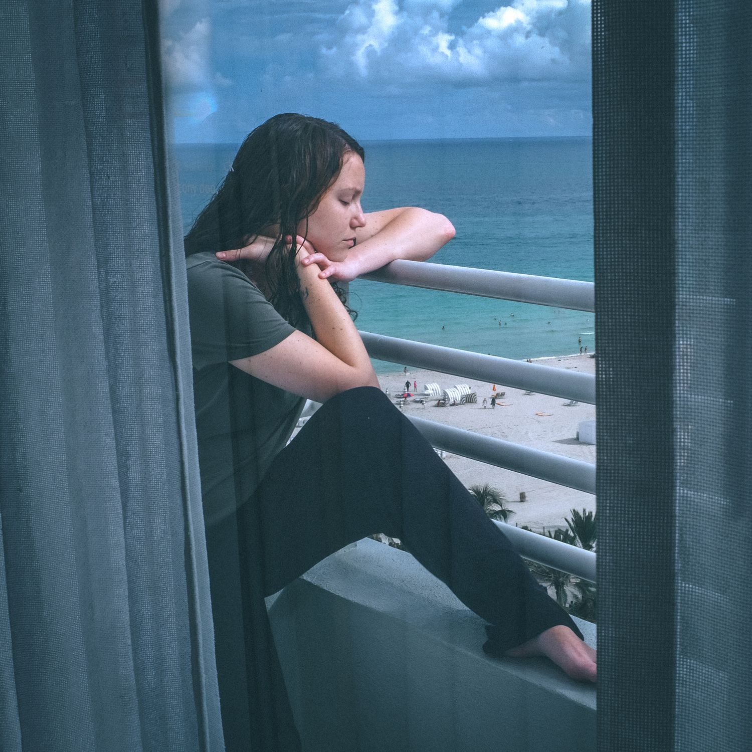 Person looking out through window curtains at a beach view with blue ocean and cloudy sky.