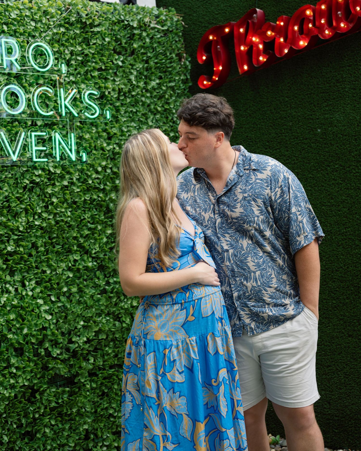 A couple in coordinating blue outfits share an intimate moment against a green hedge wall with red lights.