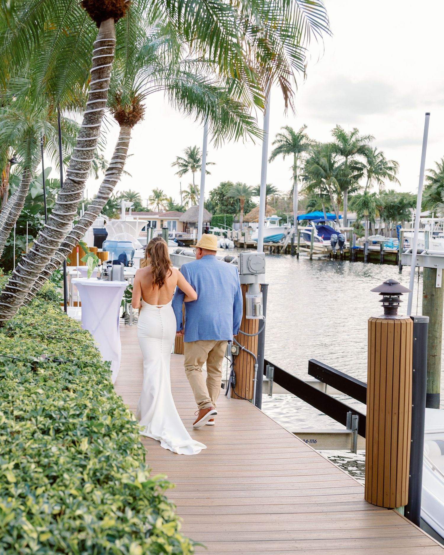 A couple walks along a tropical marina boardwalk lined with palm trees and boats.