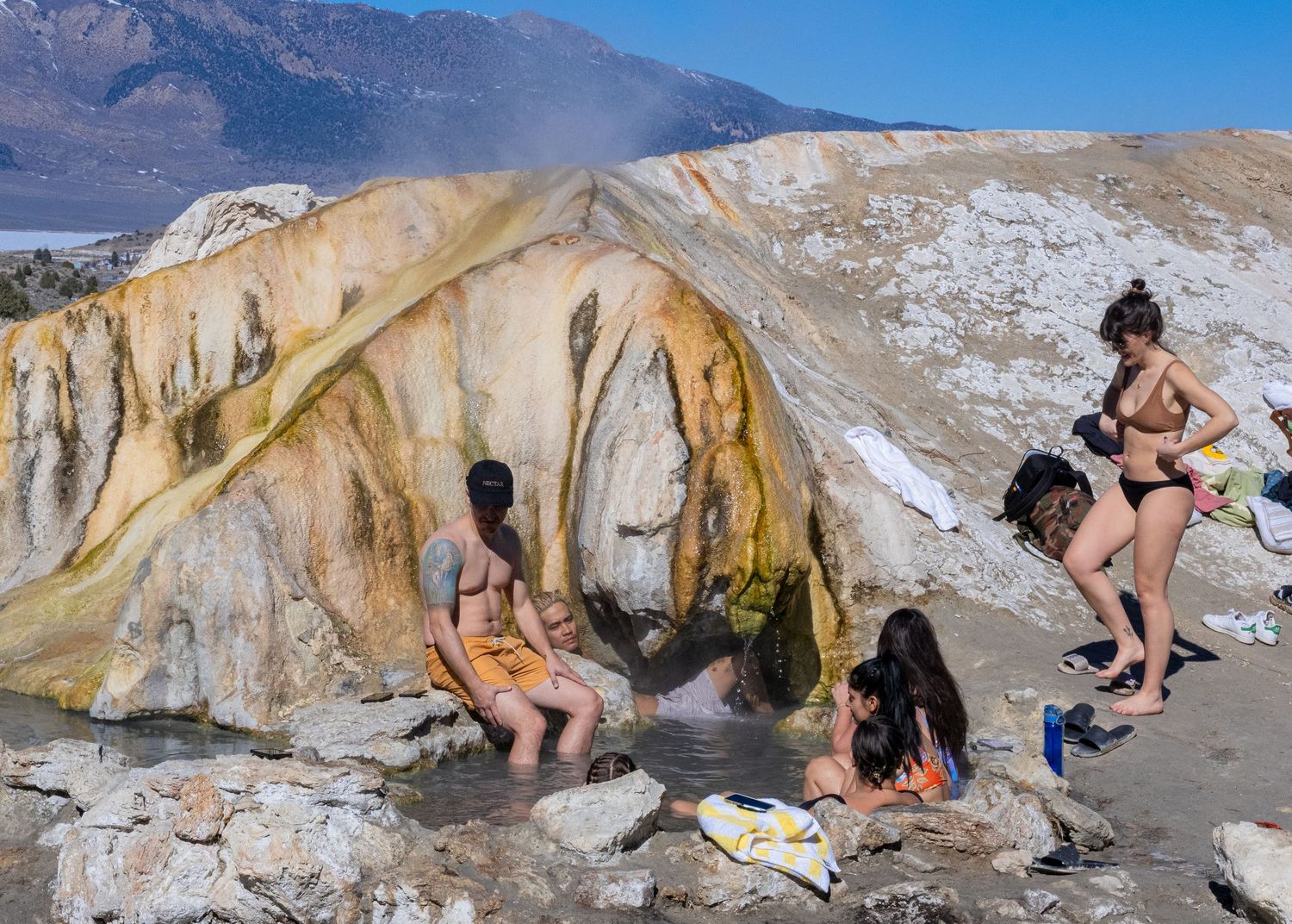 People exploring and relaxing at colorful mineral hot springs in a mountainous landscape.