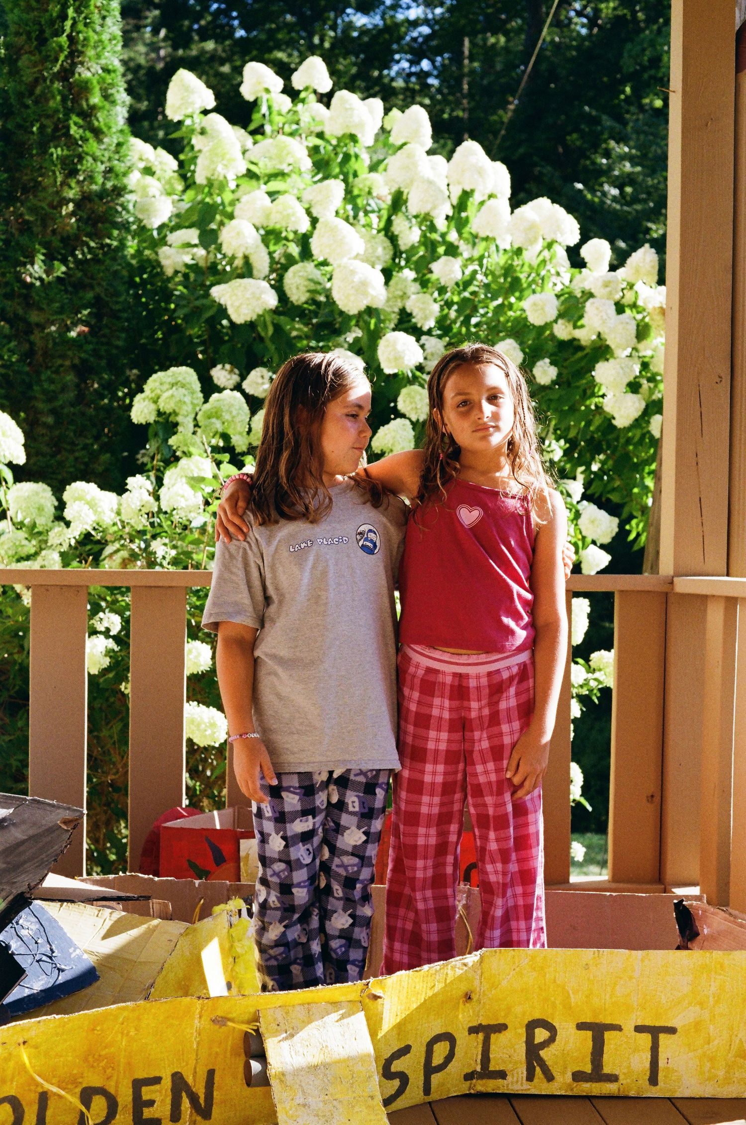 Two girls in pajamas pose on a porch with white hydrangea flowers blooming behind them on a sunny day.