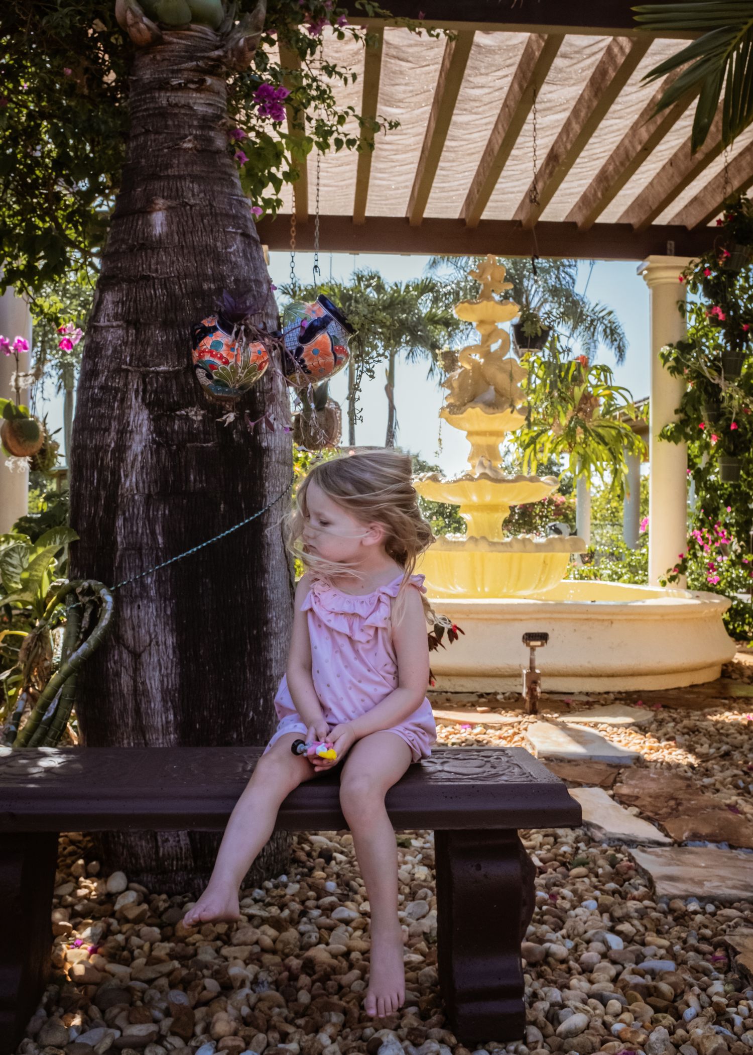 Child sits on wooden bench under a shaded pergola surrounded by tropical plants and palm trees in garden setting.