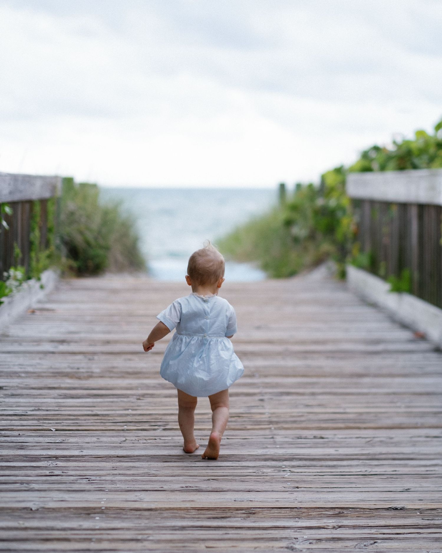 A small child in a gray shirt crawls along a wooden boardwalk path leading to the beach.
