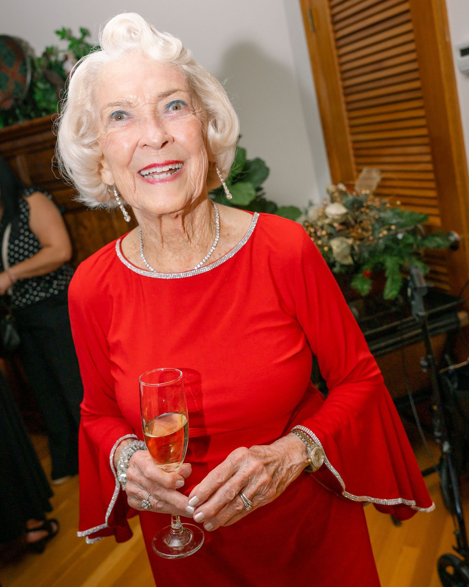 Elegant senior wearing a festive red dress holds a champagne flute while smiling warmly at a holiday gathering.