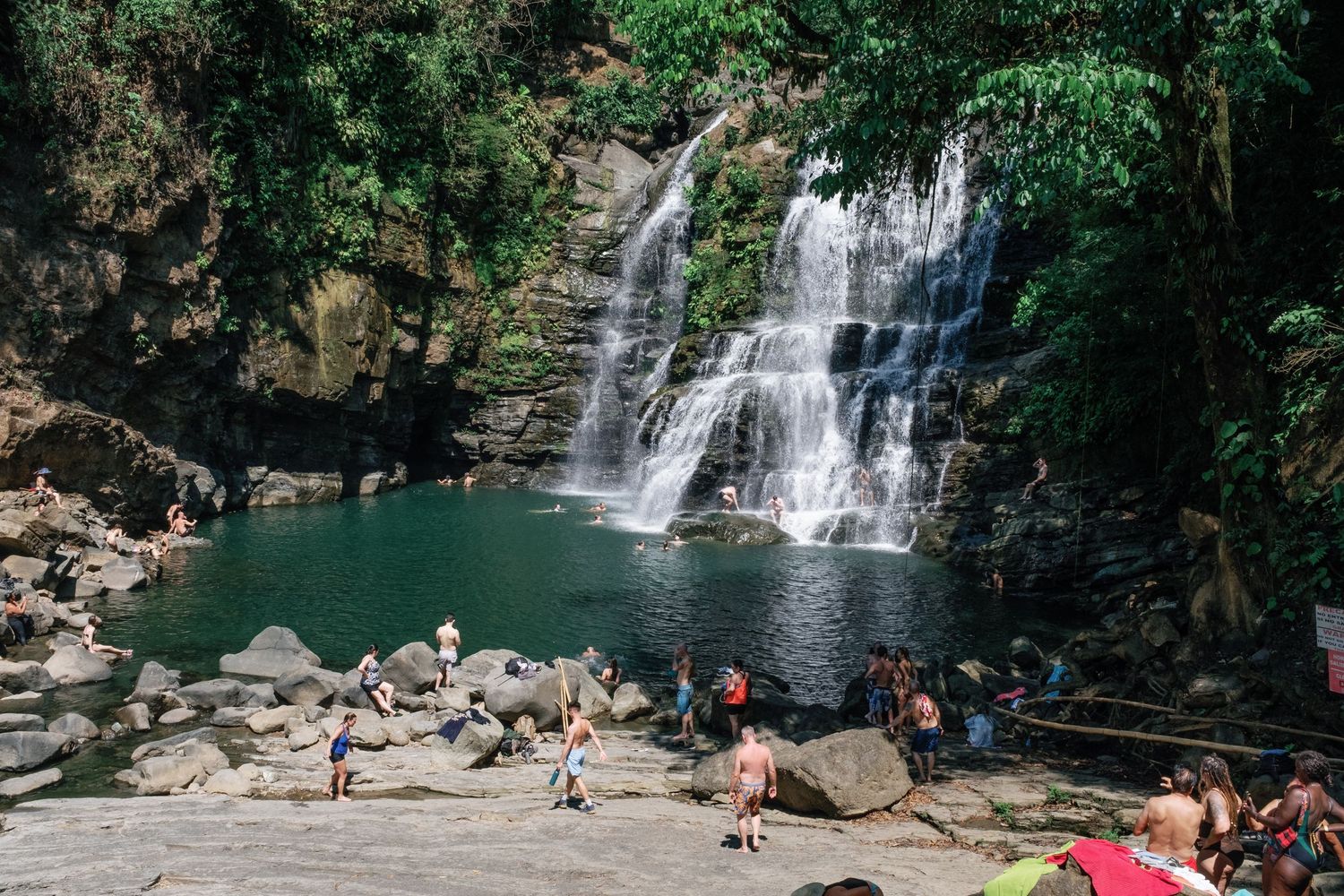 A scenic waterfall cascades into a turquoise pool where visitors relax on surrounding rocks.