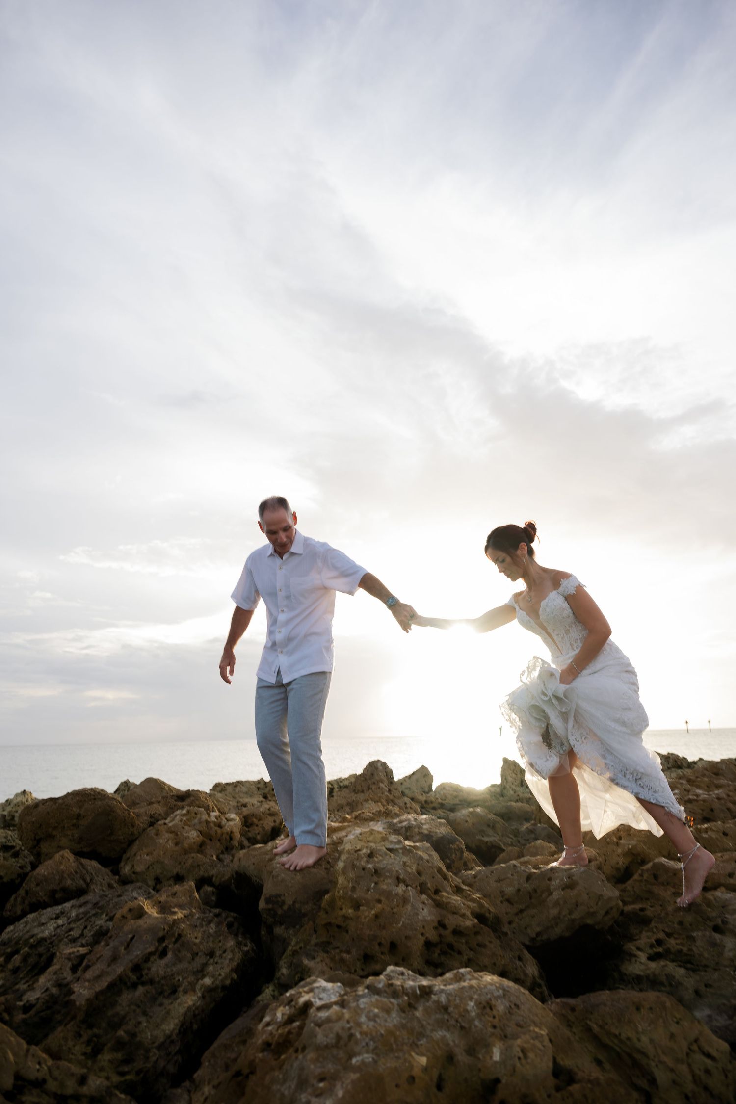 Couple in white clothing playfully climb over rocky coastal terrain at sunset.