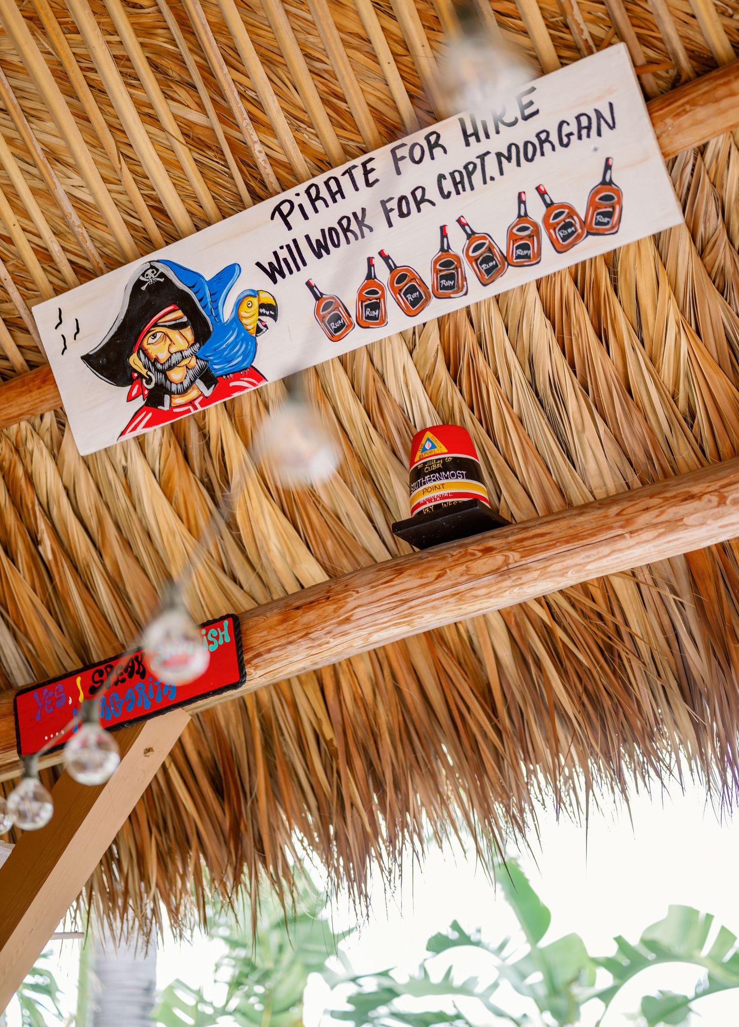 Decorative tiki bar sign with tropical themed ornaments hanging from thatched roof.