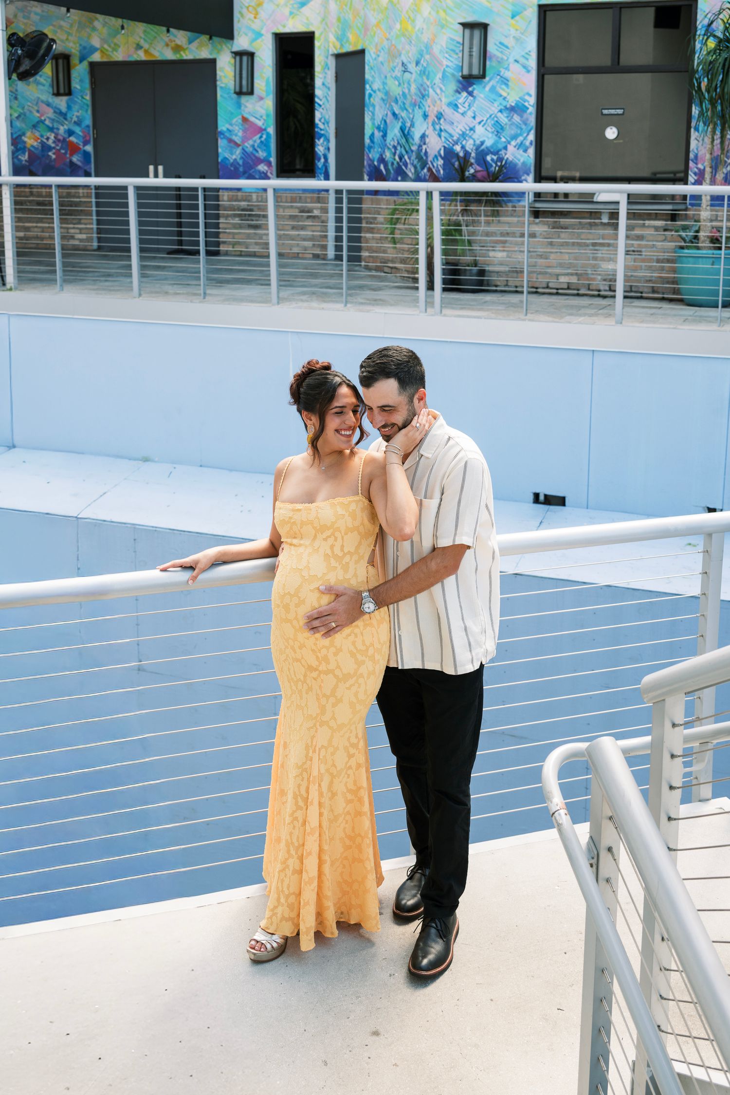 A couple in formal attire shares a romantic moment on a modern white balcony with metal railings.