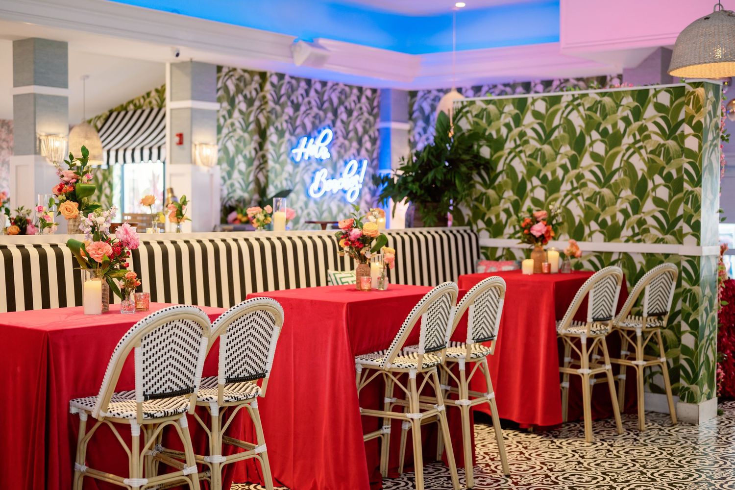 A modern restaurant interior with red banquettes and rattan chairs under pink and blue lighting.