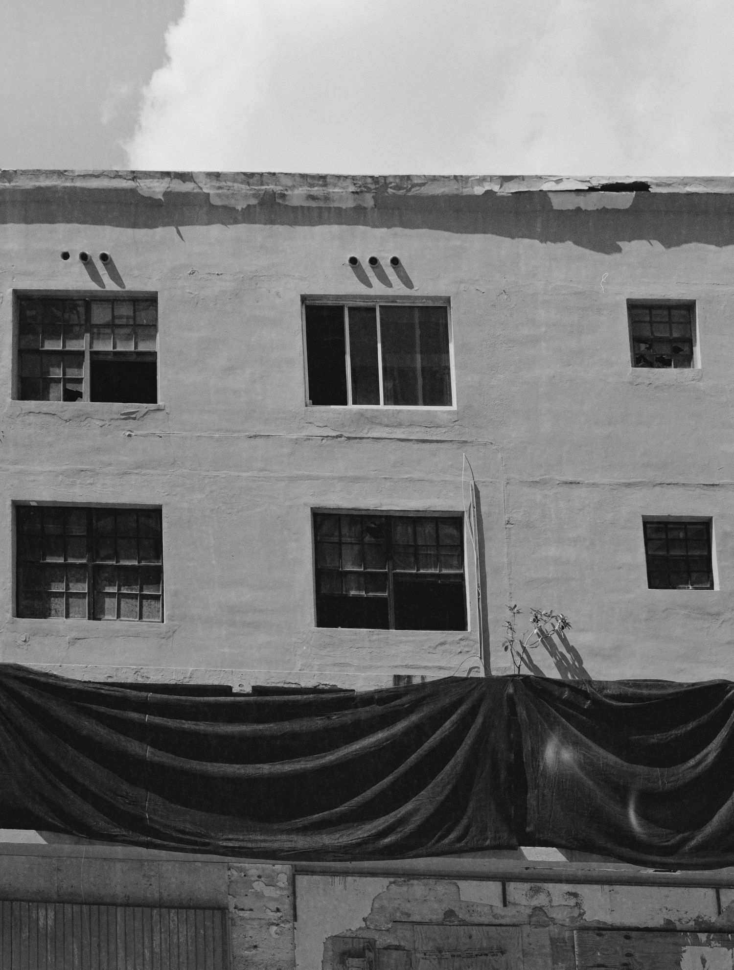 Black and white photo of an abandoned industrial building with broken windows and dark tarp draped across the facade.