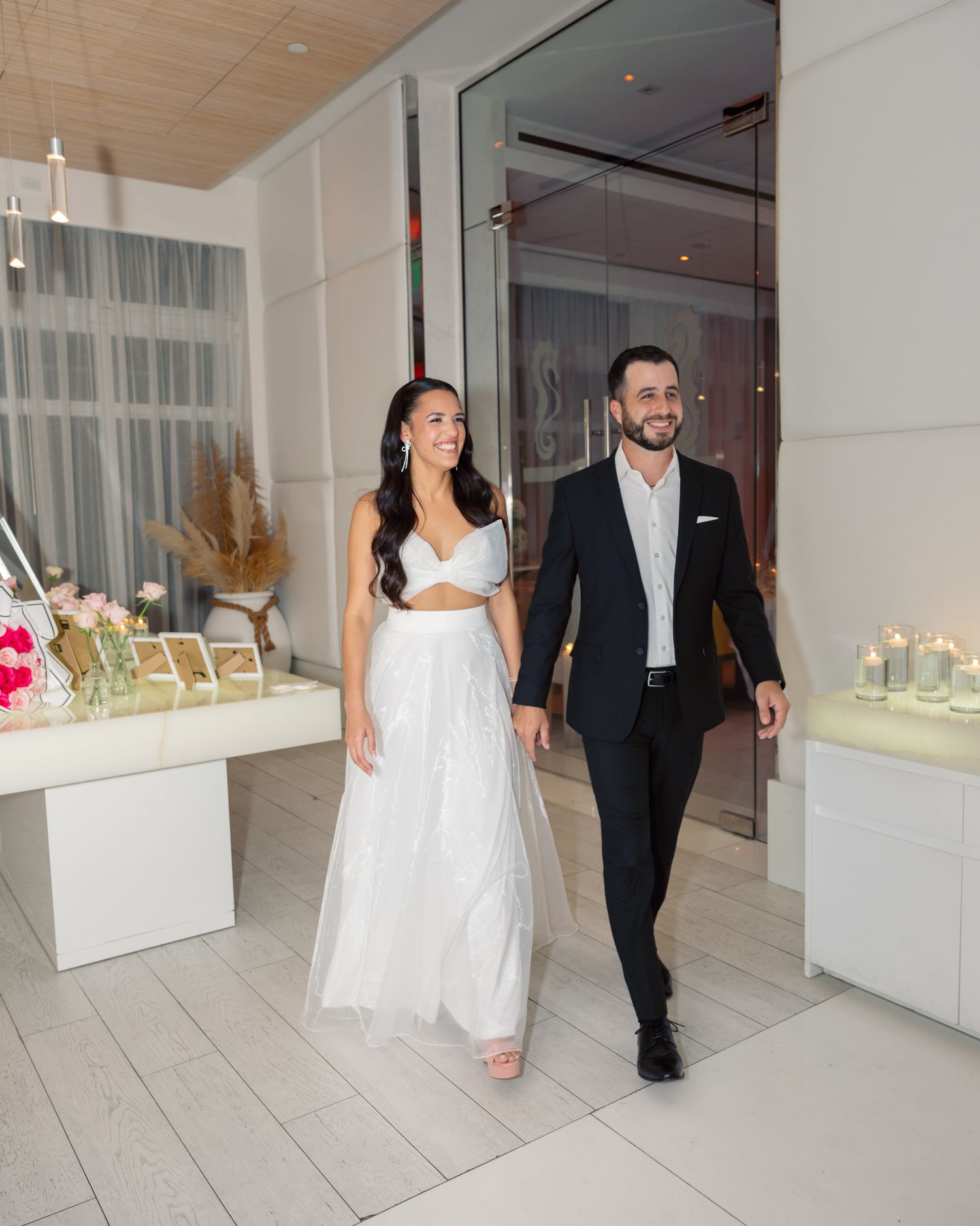 A couple in formal wedding attire walks together through a modern gallery space with white walls and display cases.