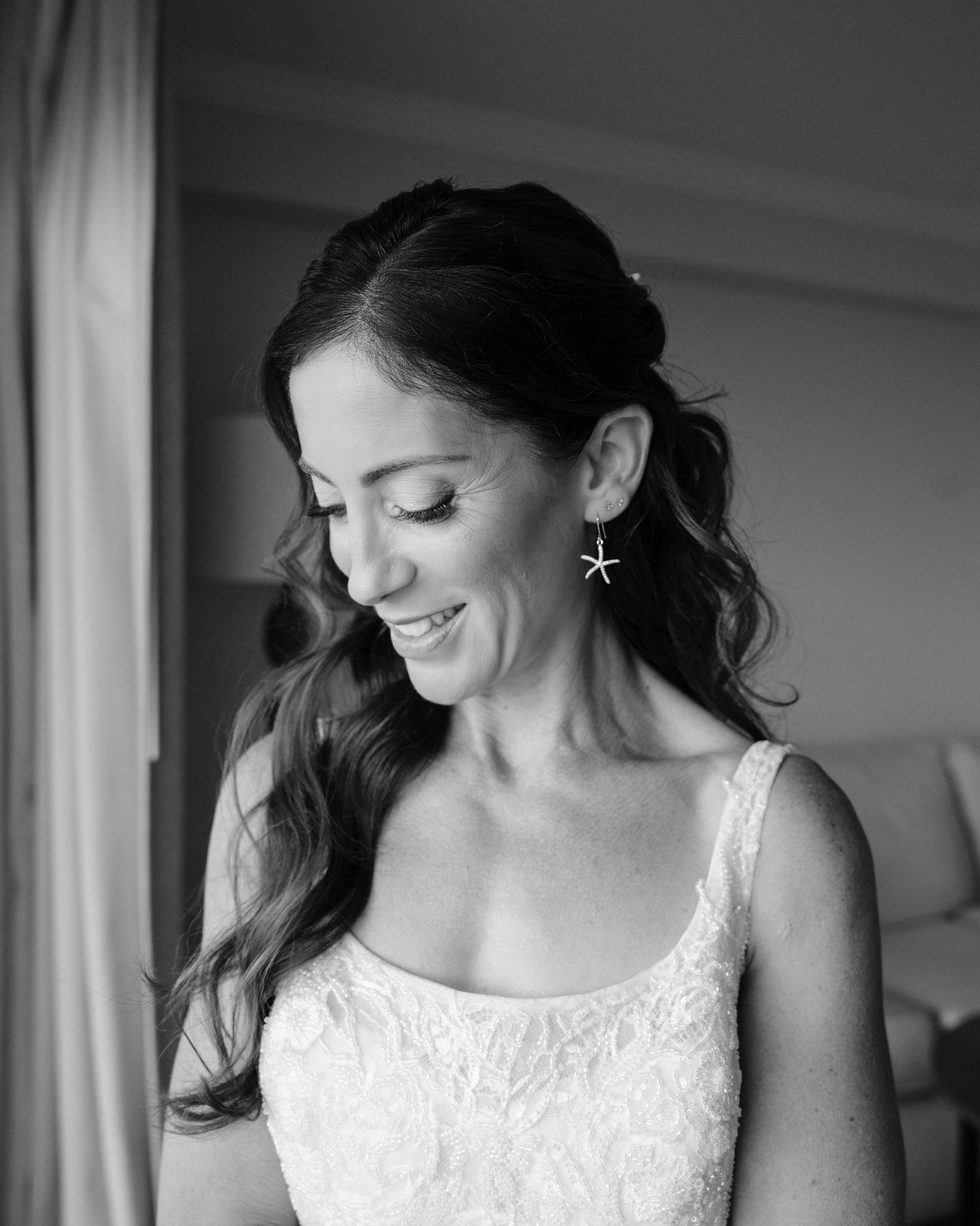 A bride in a white lace wedding dress smiles while getting ready in a black and white photograph.