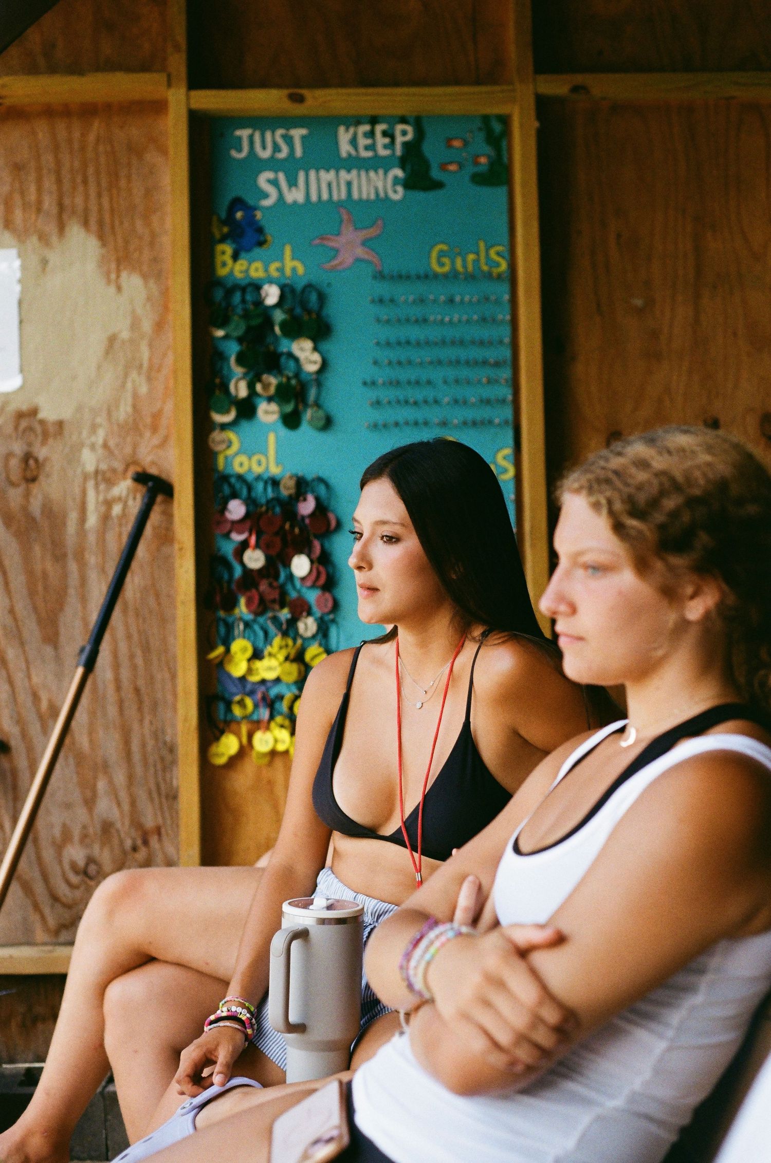 Two young women sit near a colorful camp sign reading Just Keep Swimming at an outdoor beach pool area.
