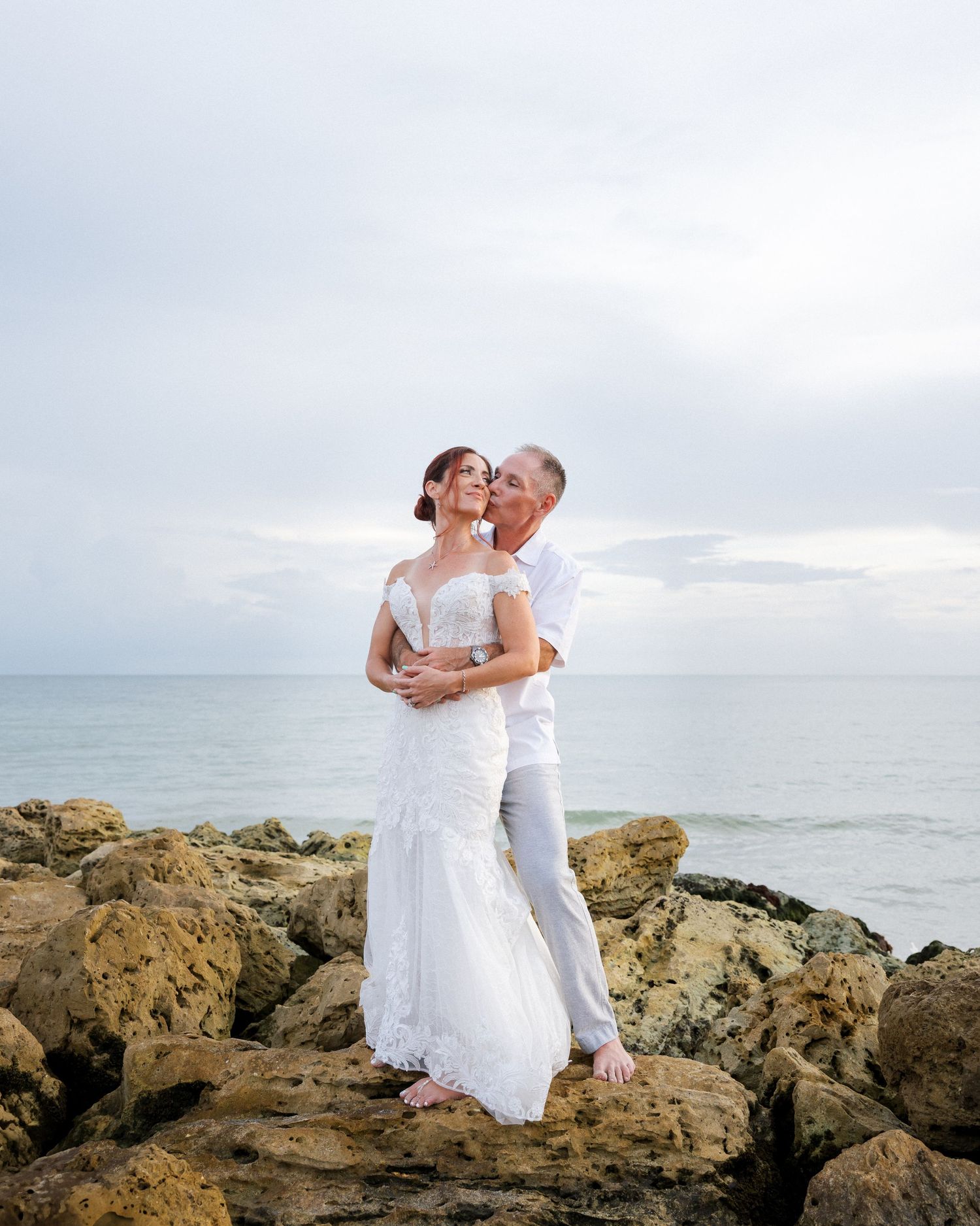 A couple in white attire embraces on rocky coastal cliffs overlooking the ocean during a romantic photoshoot.