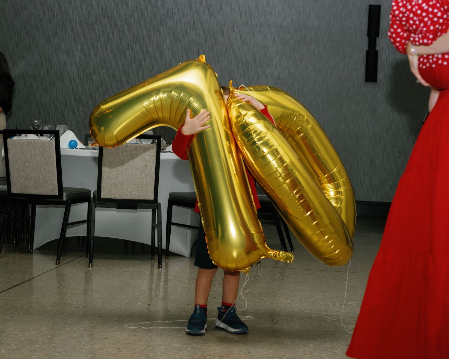 A person in a red dress stands next to large gold number balloons at an indoor event.