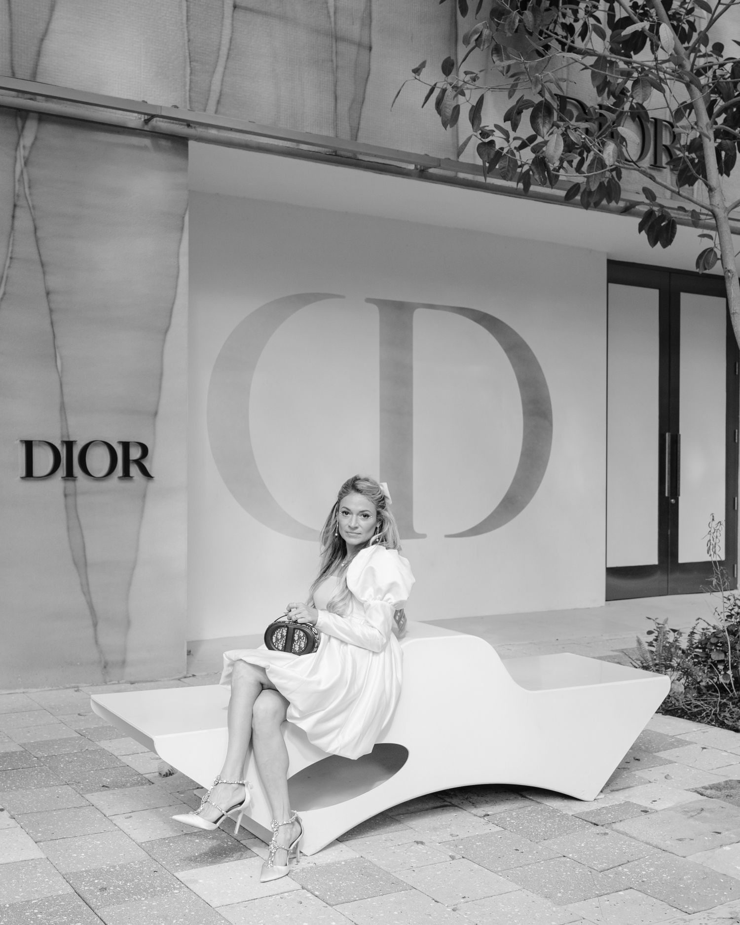 A fashion model in a flowing white dress poses elegantly against a wall with the DIOR logo in a black and white photo.