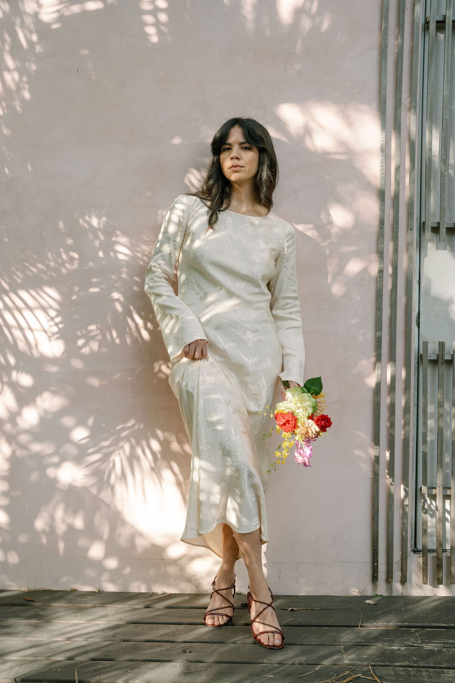 Woman in white satin midi dress holding colorful flowers, standing against a pink wall with palm tree shadows.
