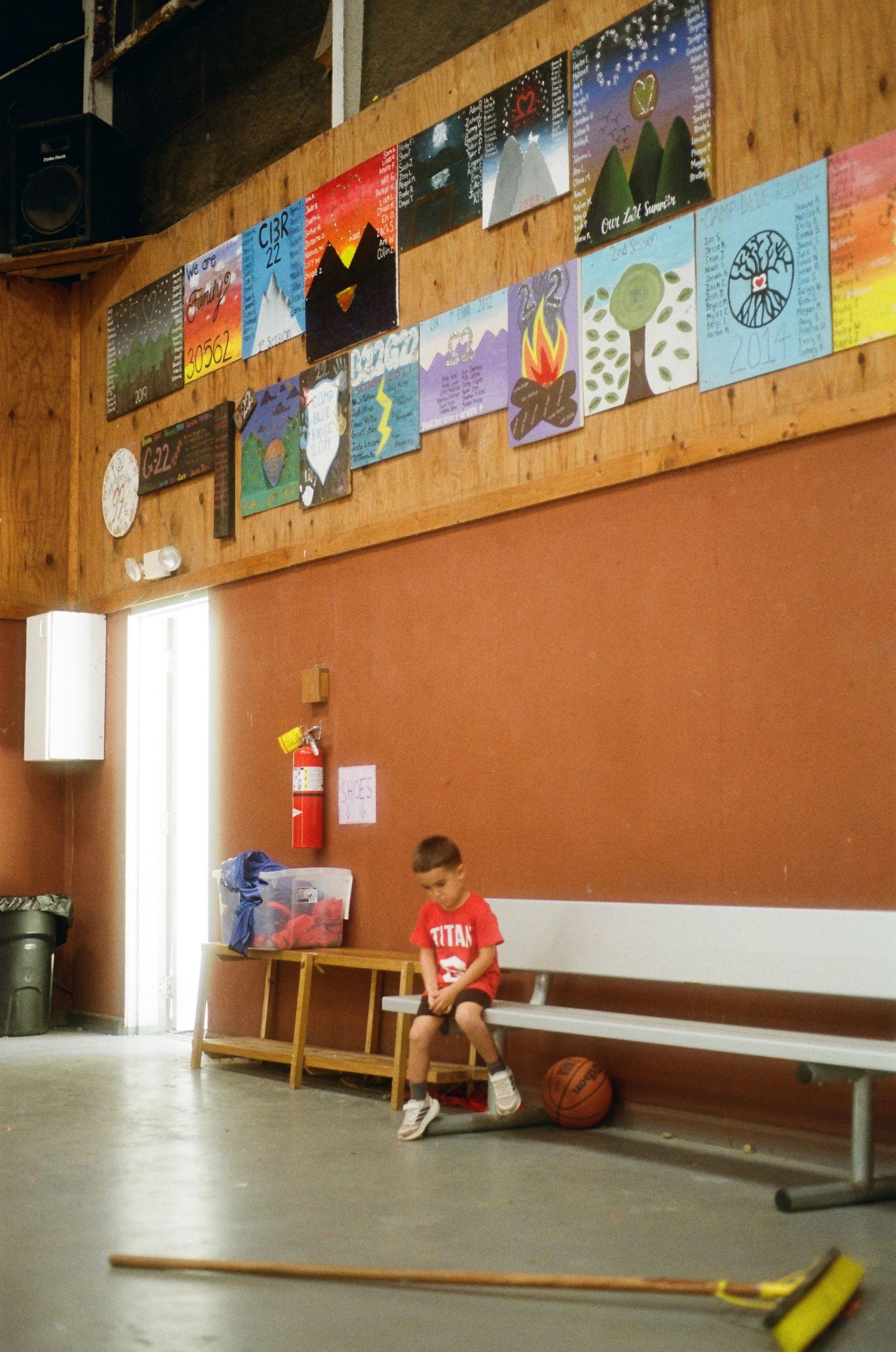 Young boy sitting alone on a bench in a school gymnasium with colorful artwork displayed on the wall above.