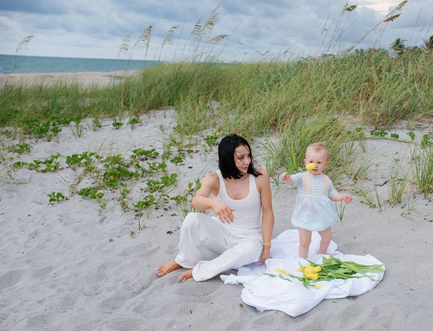 A woman in a white dress lounges on a sandy beach with sea grass and ocean waves in the background.
