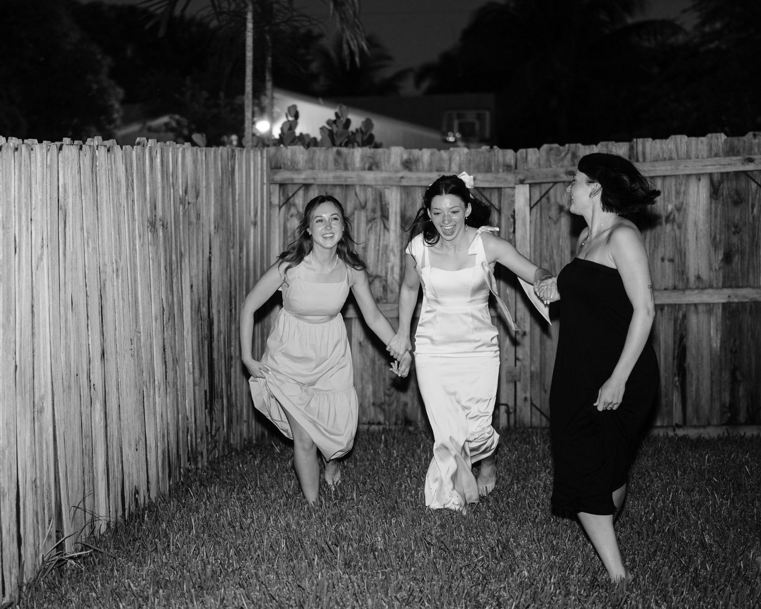 Three women in dresses run joyfully along a wooden fence in a black and white photograph.