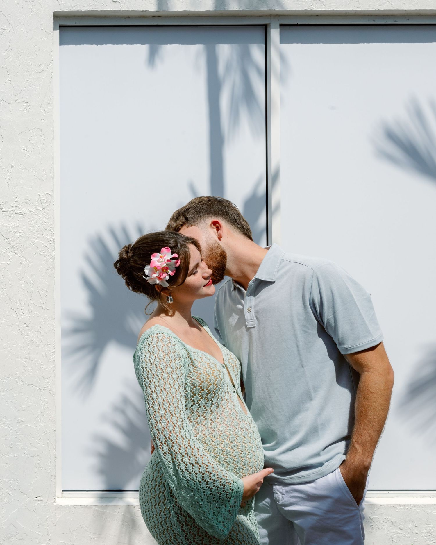 A couple shares an intimate moment against a white wall with palm tree shadows creating a romantic ambiance.