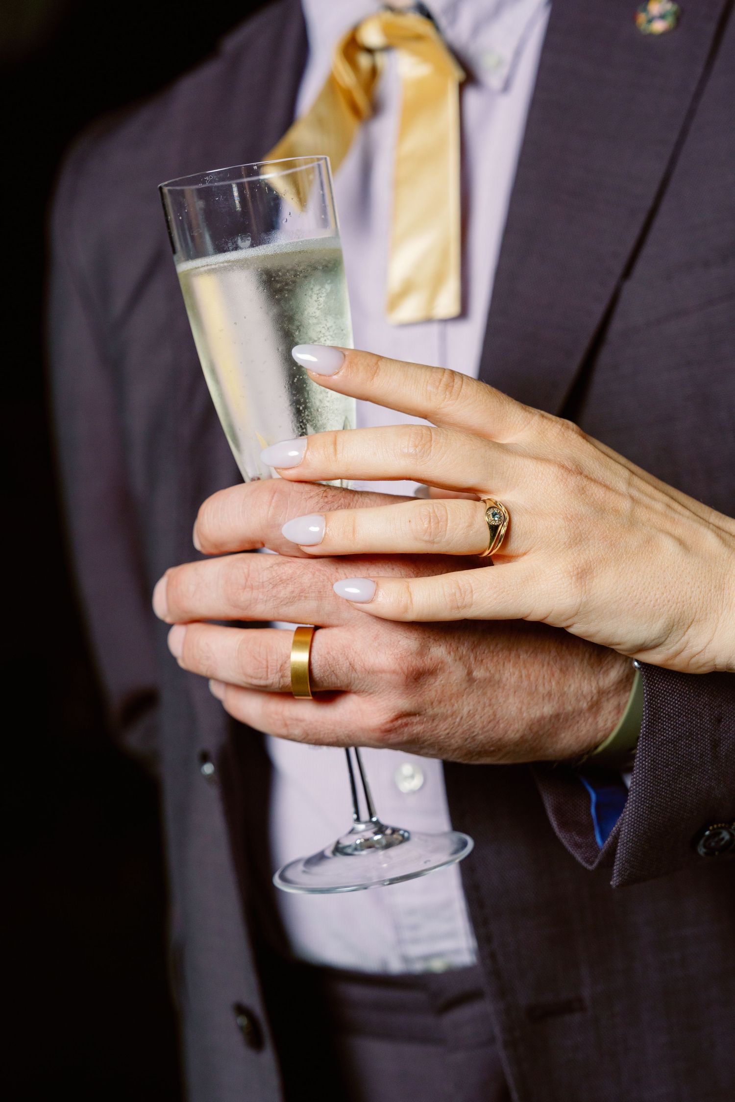 Close-up of hands holding champagne glasses showing wedding rings and formal attire.