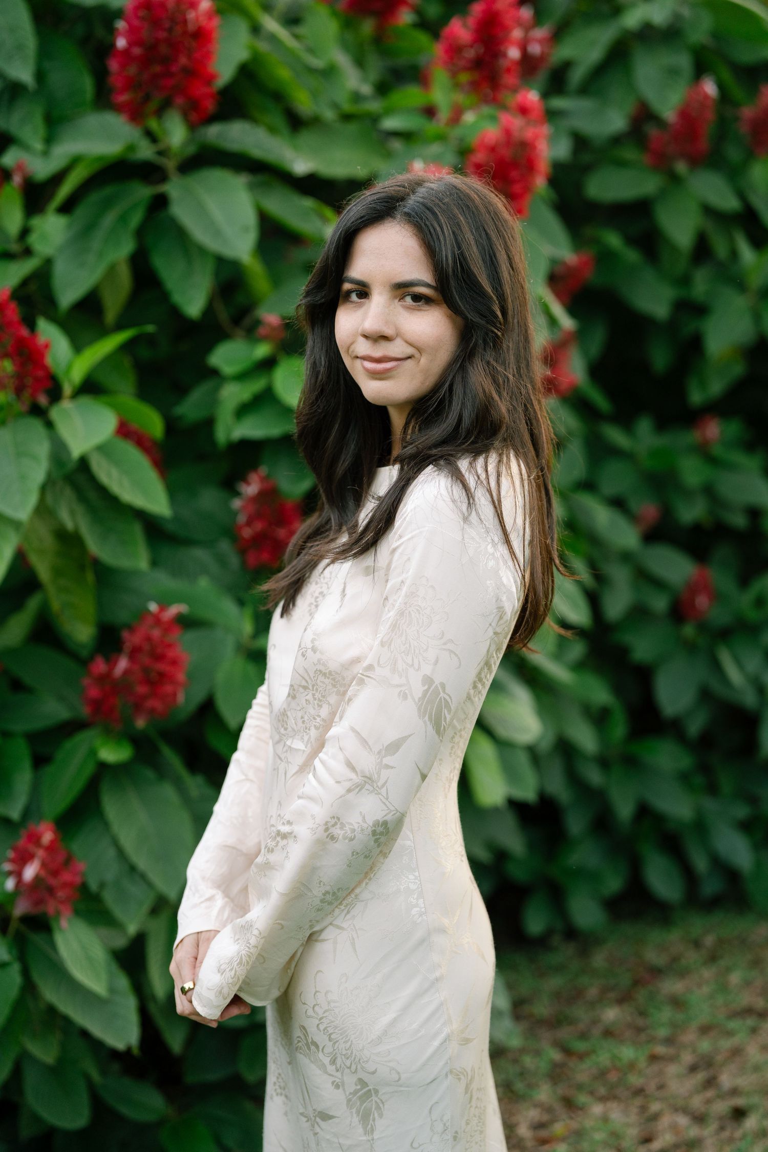 A person in a white lace dress poses in front of vibrant red flowering bushes in a garden setting.